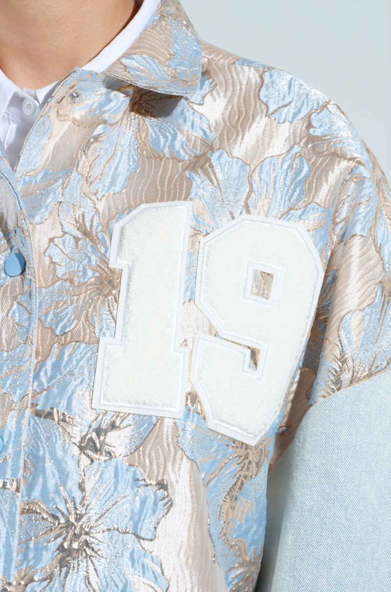 Close-up of a person wearing the ALIYA BROCADE BOMBER, showcasing its metallic floral brocade pattern in beige and blue. The jacket features a large white "19" on the chest, light denim sleeves, and a crisp white shirt collar visible underneath.