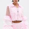 A person wears the ultra-kawaii CANDY LACE TRIM WINDBREAKER in light pink with sheer ruffled sleeves, white lace trim, and bow details. They have short dark hair, hoop earrings, a torso tattoo, and stand against a plain white background.
