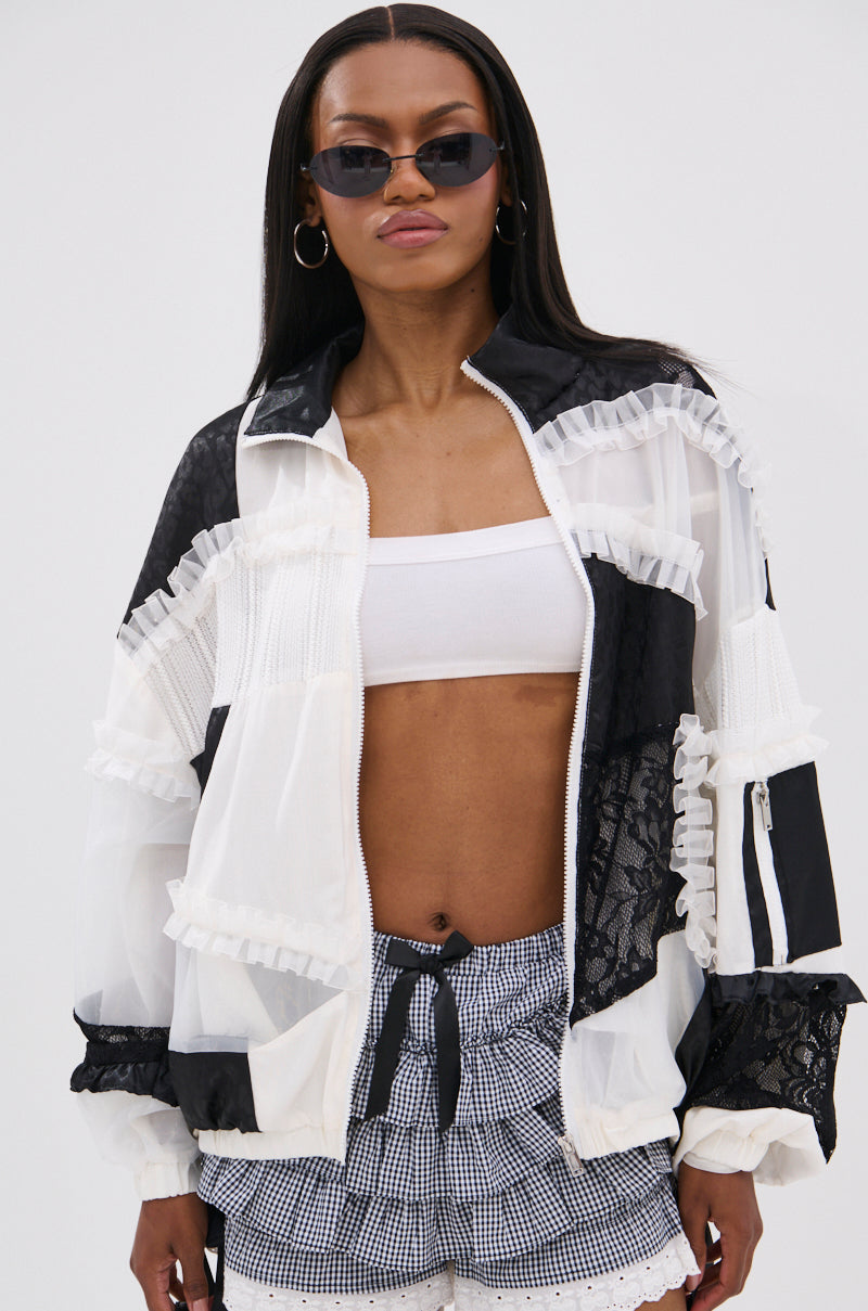 A woman stands facing the camera against a plain background, wearing dark sunglasses, hoop earrings, and the BLACK DAHLIA PATCHWORK BOMBER over a white bandeau top and checkered shorts.