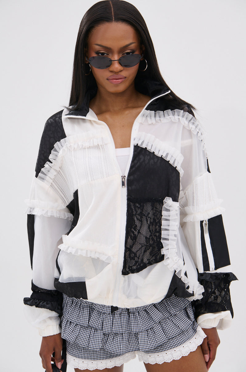 A woman in black sunglasses and the BLACK DAHLIA PATCHWORK BOMBER, featuring lace and ruffle details, pairs it with a white top and black-and-white checkered shorts as she stands against a plain white background.