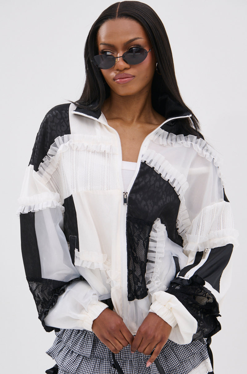 A woman wears sunglasses and the BLACK DAHLIA PATCHWORK BOMBER, featuring sheer mesh and ruffled details, standing confidently against a plain background in this bold statement jacket.