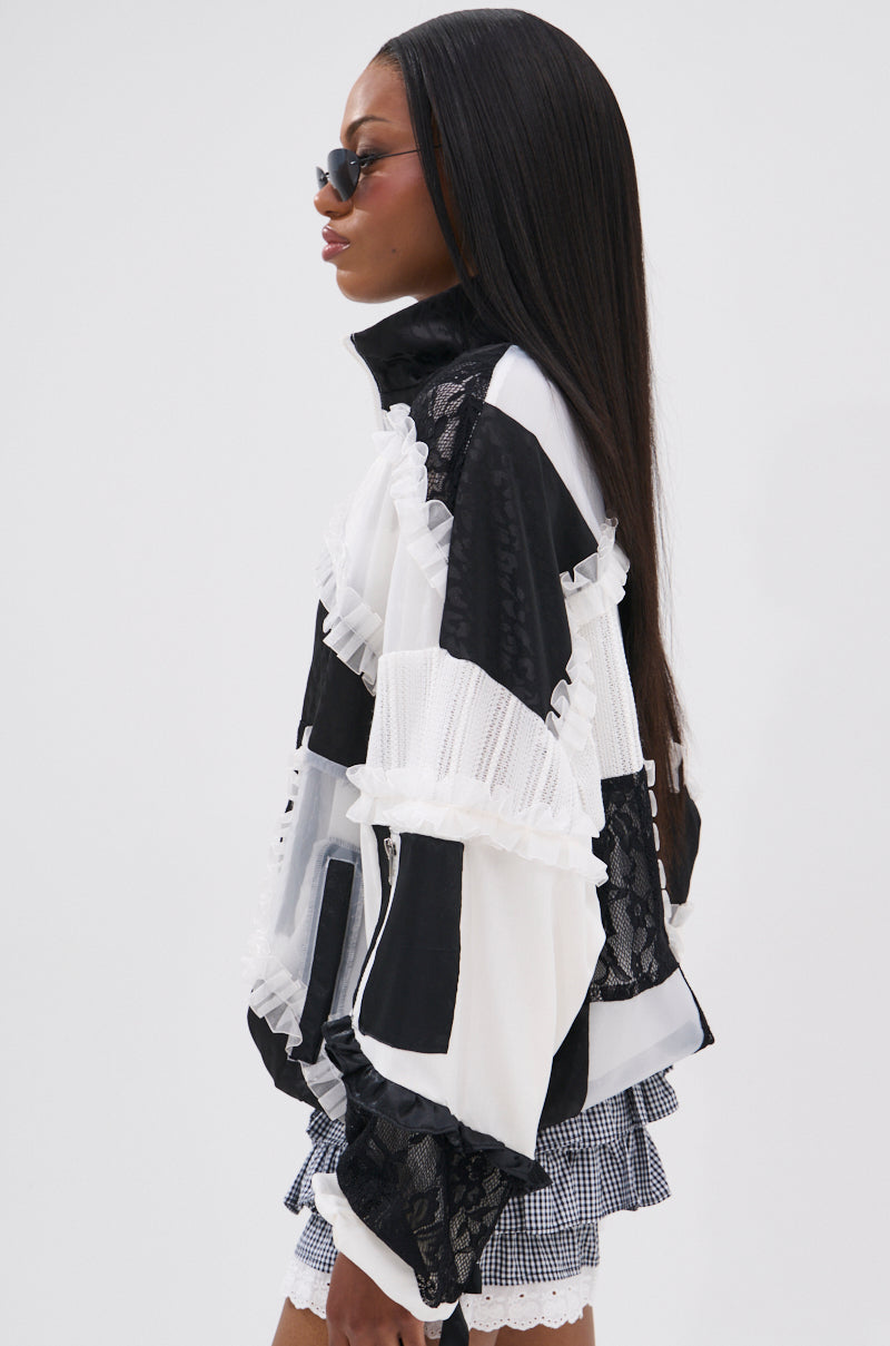 A woman with long straight hair and sunglasses models the BLACK DAHLIA PATCHWORK BOMBER, featuring lace details, paired with plaid shorts. She stands in profile against a plain white background.