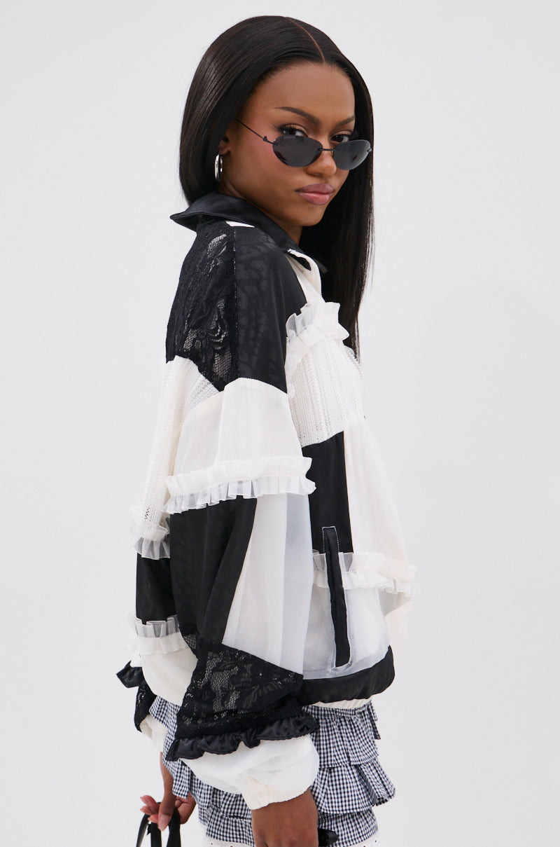 A woman with long straight hair wears black sunglasses, holds a small black purse, and poses against a white background in the BLACK DAHLIA PATCHWORK BOMBER featuring black and white ruffles, lace details, and a checkered skirt.