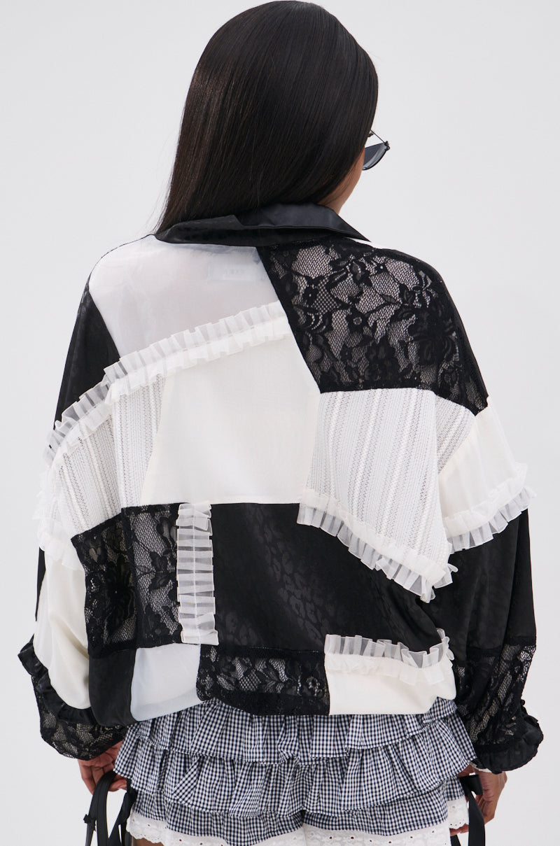 A person with long dark hair, seen from behind, wears the BLACK DAHLIA PATCHWORK BOMBER—featuring lace and ruffled accents—styled with black and white checkered shorts.