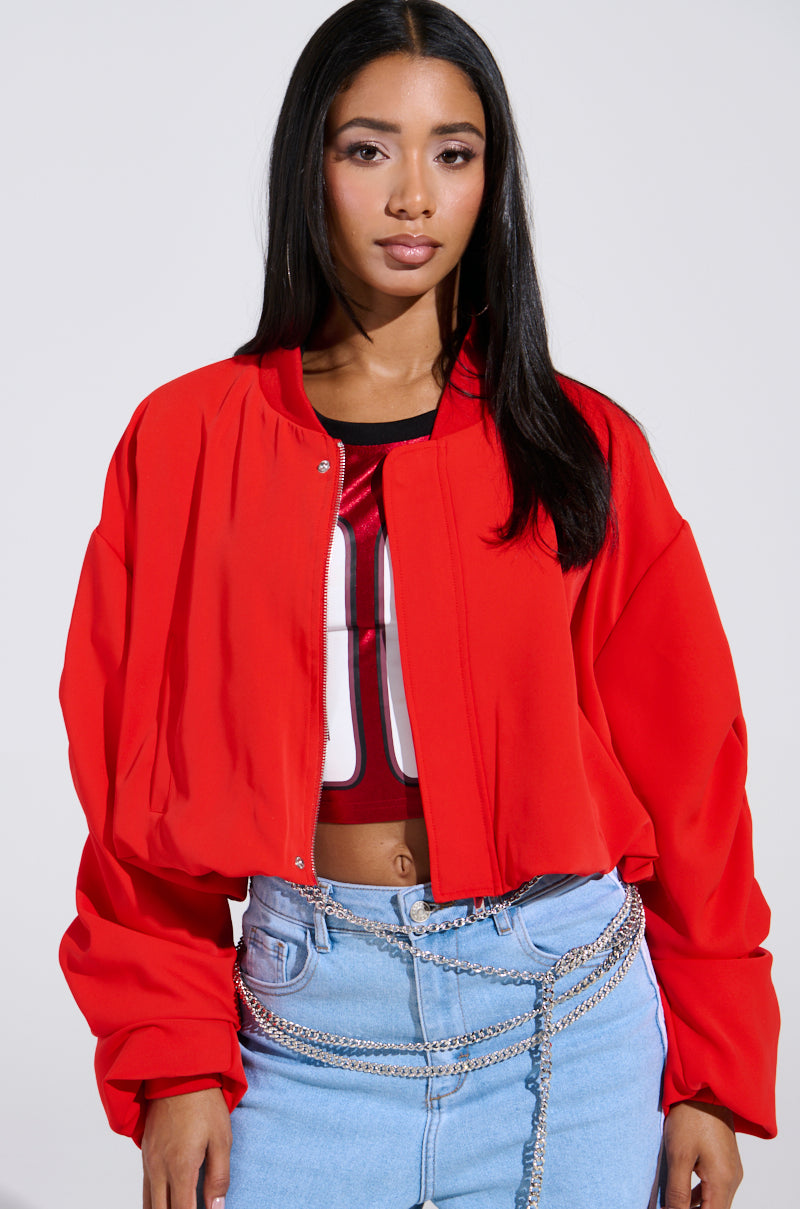 A person with long dark hair stands confidently in the TREY Ruched Sleeve Bomber in red over a red top with white numbers. They pair it with light blue jeans and a silver chain belt, set against a simple backdrop, exuding cherry bomb vibes.