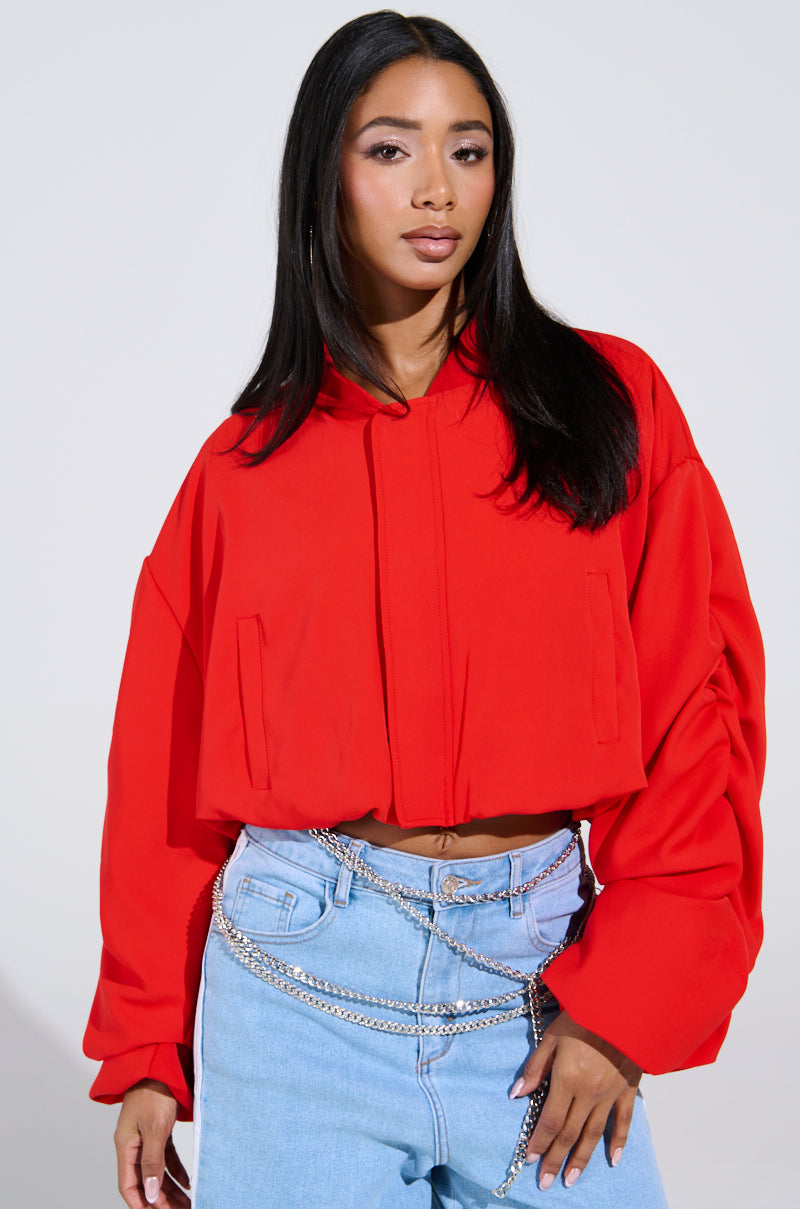 A person with long dark hair confidently poses against a plain, light background wearing the TREY Ruched Sleeve Bomber in red and light blue jeans with chain details.