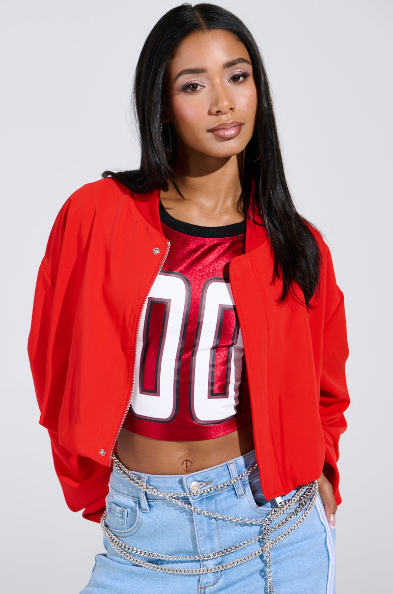 A woman with long black hair confidently showcases her style, wearing the TREY RUCHED SLEEVE BOMBER IN RED over a red top with white "00," paired with light blue jeans accented by chain accessories. The jacket's cropped fit and ruched sleeves enhance its trendy flair against a light background.
