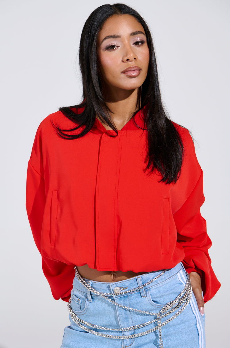 A person with long dark hair exudes cherry bomb vibes in the TREY Ruched Sleeve Bomber in Red, paired with light blue jeans and silver chain belts. The plain, light-colored background accentuates their bold look.