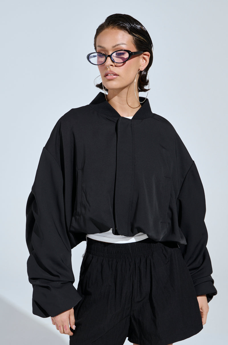 Wearing the TREY RUCHED SLEEVE BOMBER over a white top with black shorts, a person with slicked-back hair and oversized black sunglasses showcases street-style fashion against a plain light background.