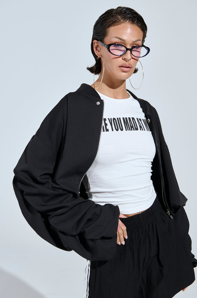 A woman stands confidently against a plain light background, embodying street style in cat-eye glasses, large hoops, a white graphic tee, black shorts, and the TREY RUCHED SLEEVE BOMBER.