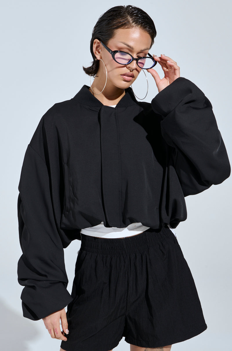 Wearing the TREY RUCHED SLEEVE BOMBER, a person in oversized black glasses and black shorts stands against a light background, looking down and adjusting their glasses in cool street-style fashion.