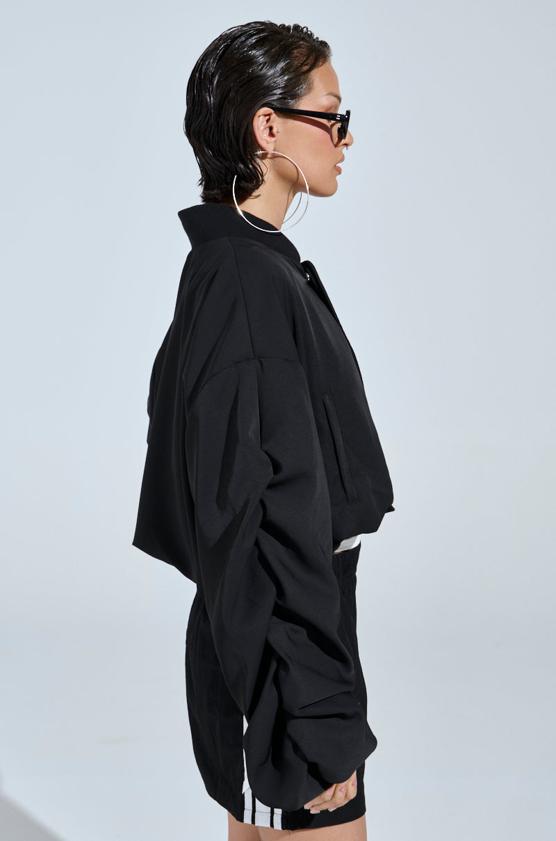 Wearing the TREY RUCHED SLEEVE BOMBER, a person with slicked-back hair, dark sunglasses, and large hoop earrings stands in profile against a plain light background, embodying effortless street-style fashion.