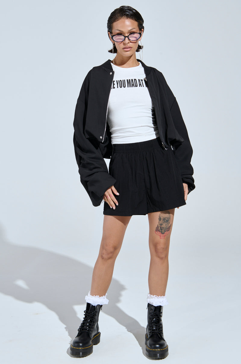 A person stands confidently in street-style fashion, wearing the TREY RUCHED SLEEVE BOMBER with black sunglasses, a white graphic T-shirt, black shorts, lace-trimmed black boots, and white frilly socks against a plain background.