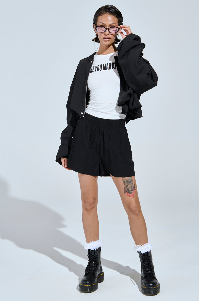 A woman exudes street-style confidence in glasses, the TREY RUCHED SLEEVE BOMBER over a white "ARE YOU MAD AT" shirt, black shorts, white socks, and black boots. She adjusts her glasses, revealing a tattoo on her right thigh.