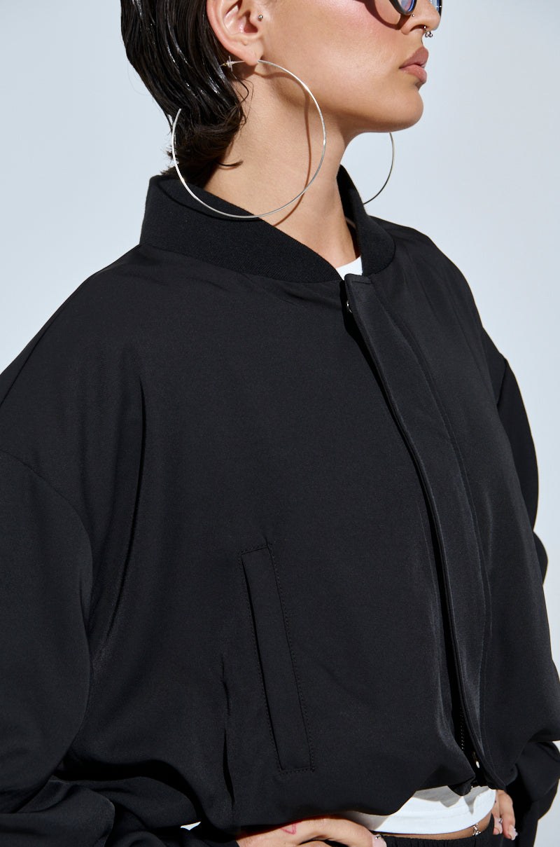 Person with short, wet-look hair wears large hoop earrings, sunglasses, and the TREY RUCHED SLEEVE BOMBER—a black jacket with ruched sleeves and high collar—showcasing bold street style. Only the lower face and upper torso are visible.