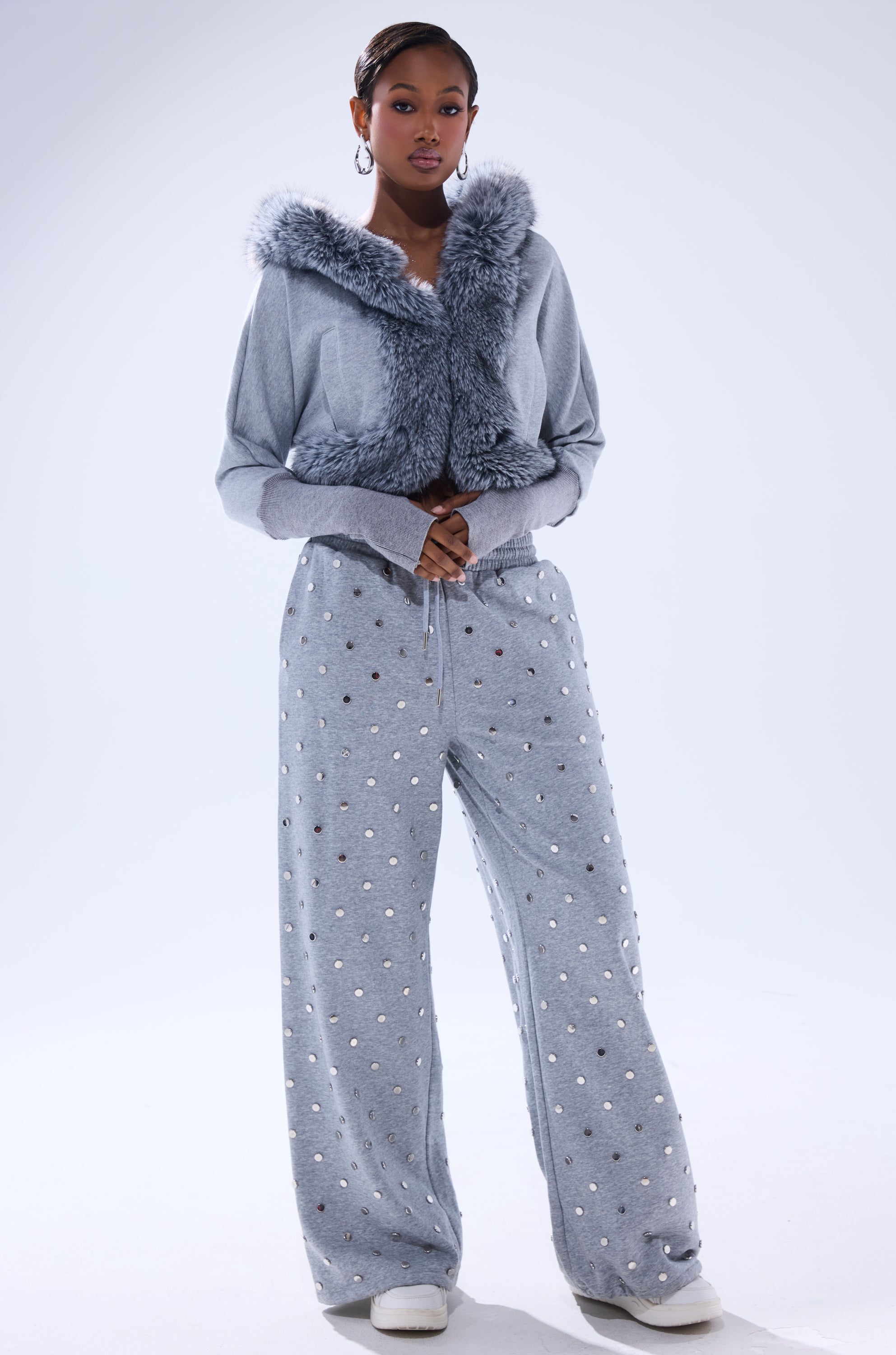 A woman wears the MOVIN DIFFERENT SWEATSHIRT WITH FUR TRIM in light grey, styled as a cropped jacket with loose grey pants featuring small white spots and white sneakers, posed against a plain light background. 9