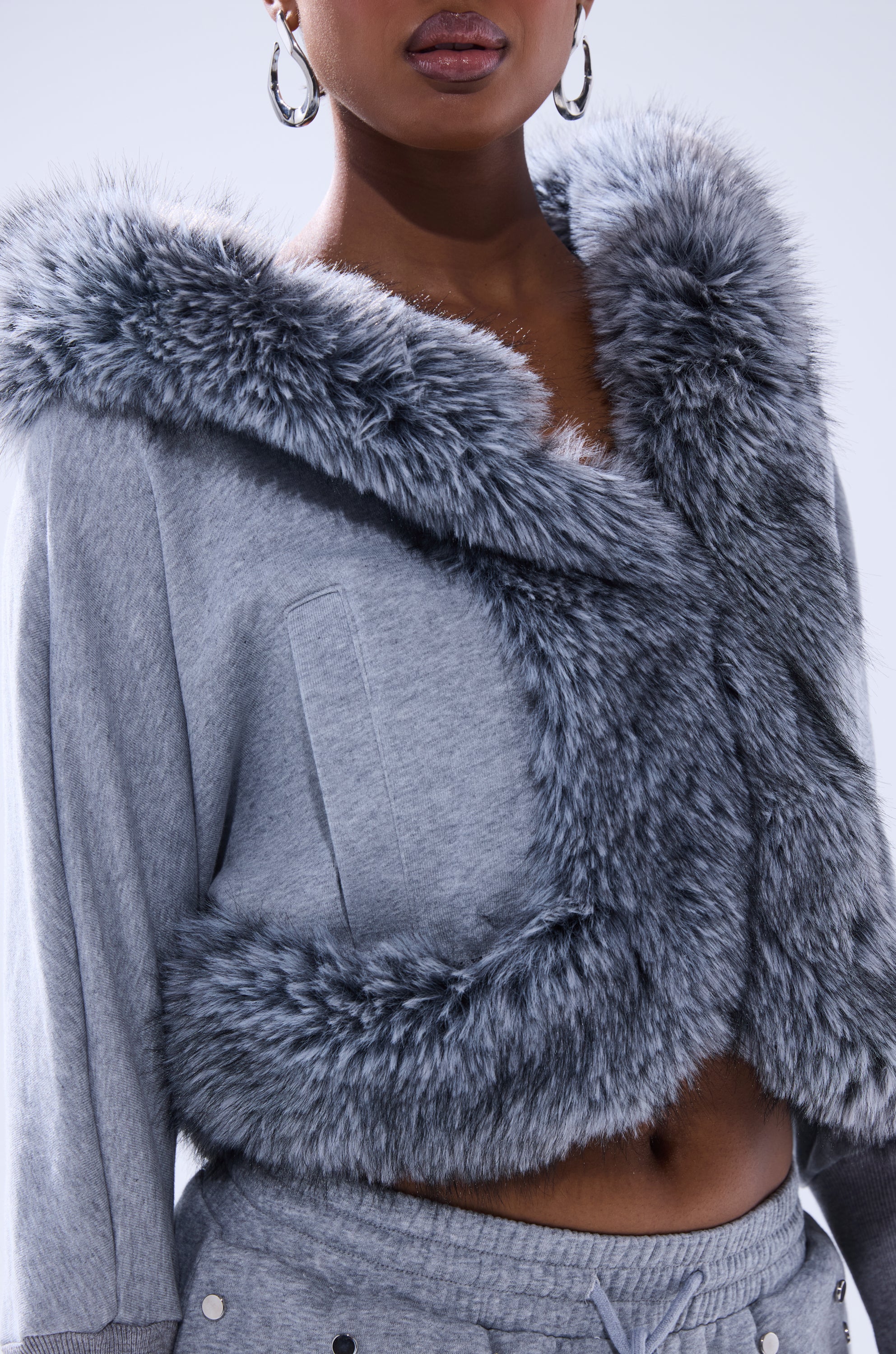 A person is shown from shoulders to waist wearing the MOVIN DIFFERENT SWEATSHIRT WITH FUR TRIM in light gray, featuring plush faux fur around the collar and front, paired with matching gray pants. 10