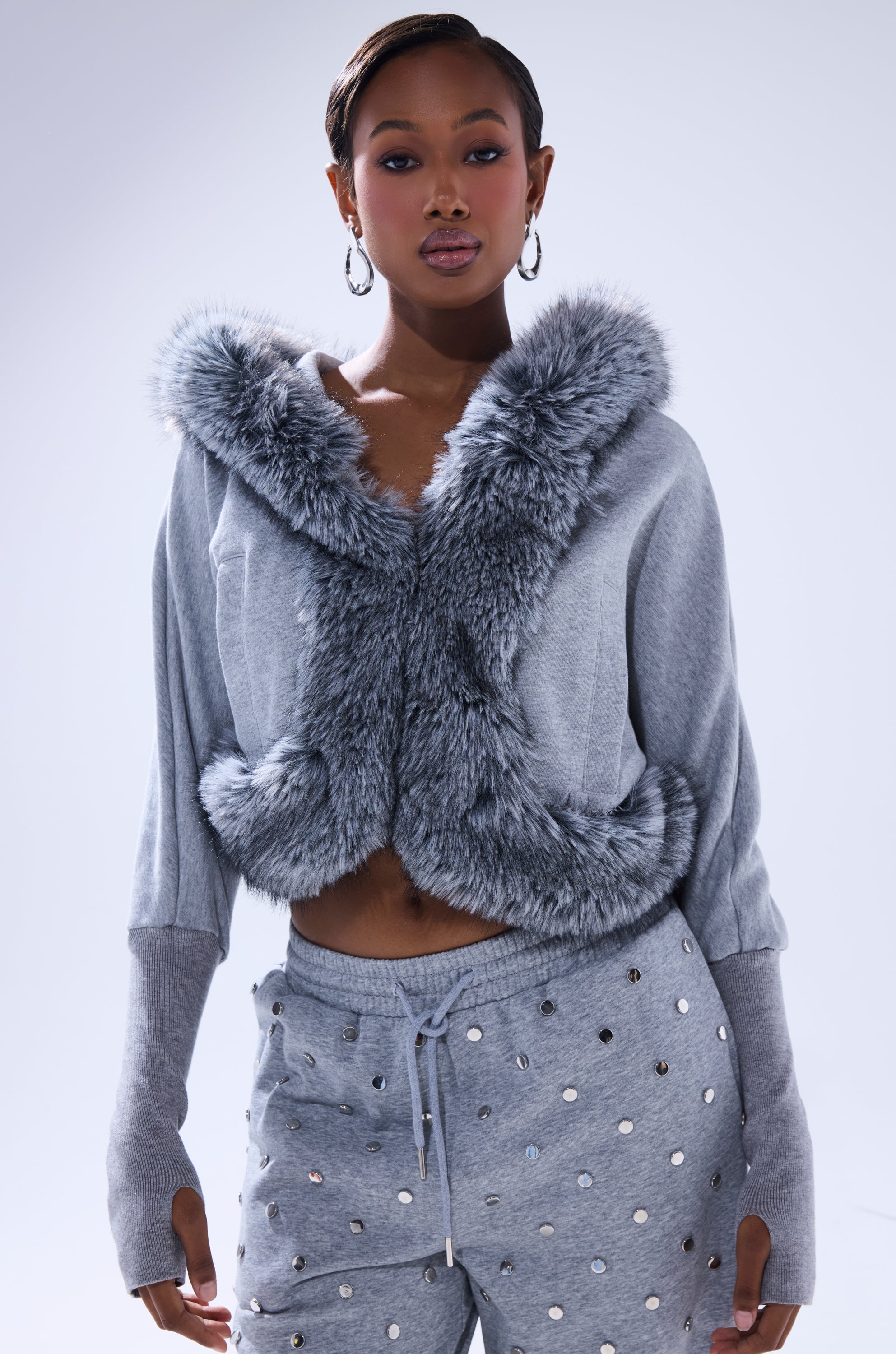 A woman stands against a light background, confidently wearing the MOVIN DIFFERENT SWEATSHIRT WITH FUR TRIM and matching gray sweatpants with metallic studs. She accessorizes with hoop earrings. 1