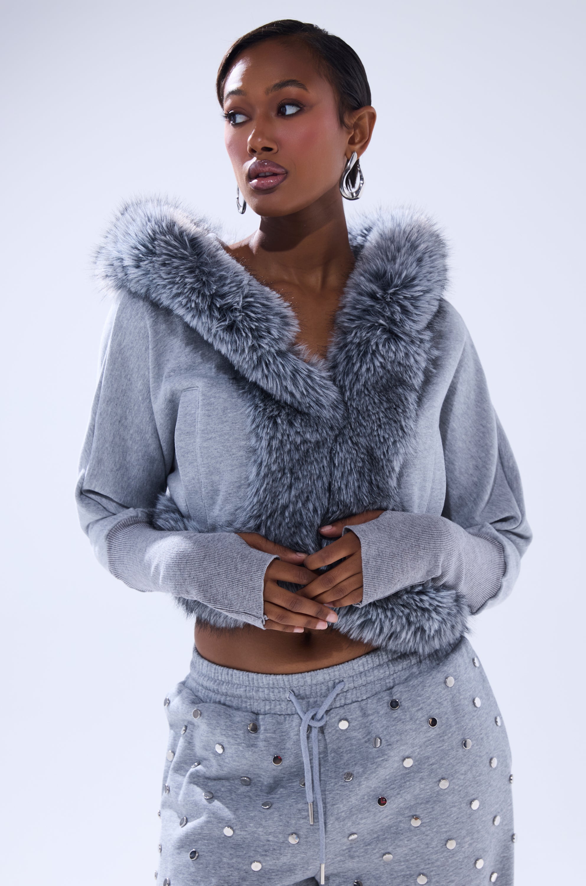 A woman wears the MOVIN DIFFERENT SWEATSHIRT WITH FUR TRIM and matching studded sweatpants, standing against a plain light background with clasped hands for an effortlessly chic look. 3