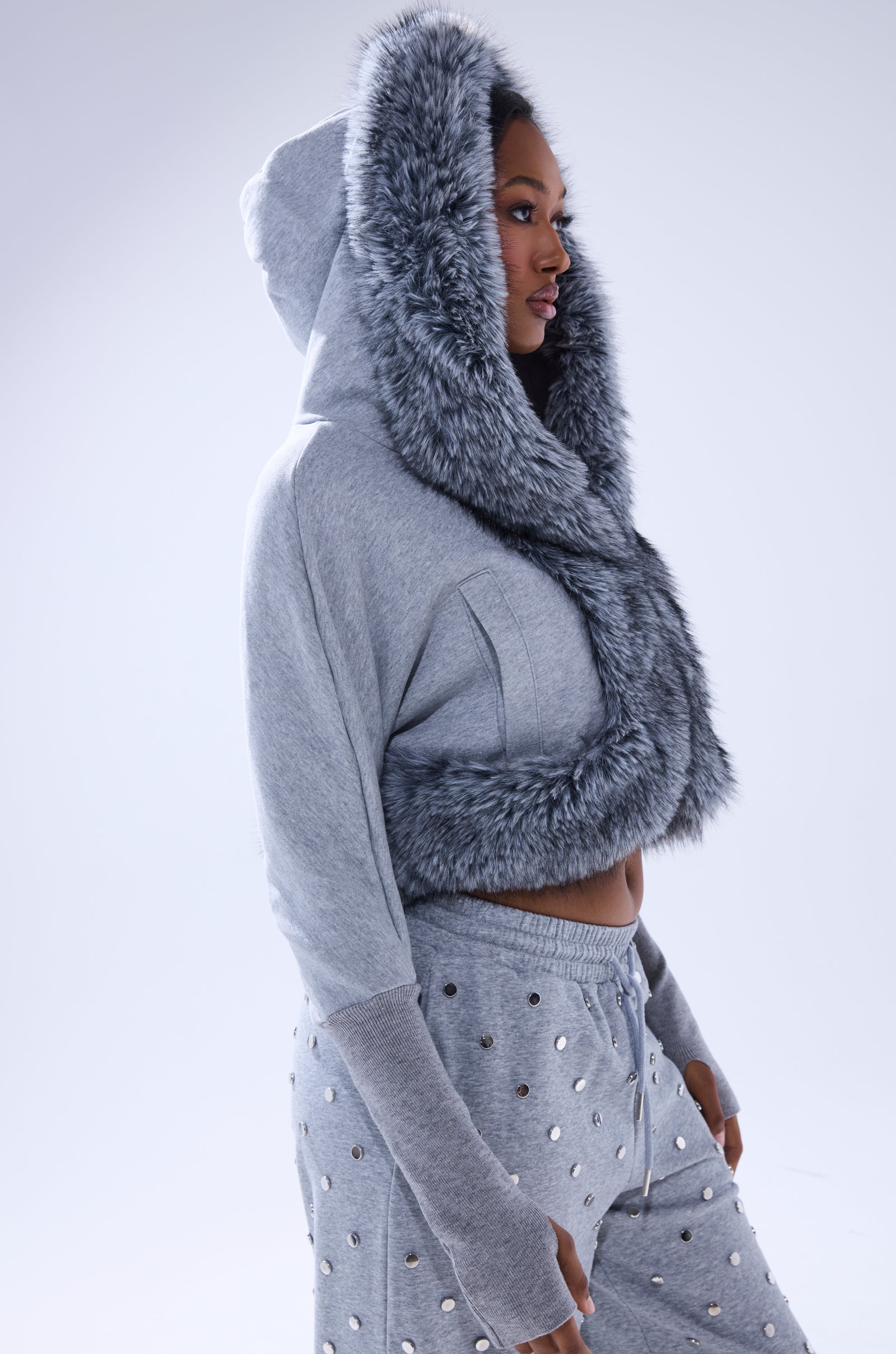A woman in profile wears the MOVIN DIFFERENT SWEATSHIRT WITH FUR TRIM in light gray, paired with matching studded gray pants. The thick faux fur-edged hood exudes luxury. The plain, light-colored background highlights the outfit. 4