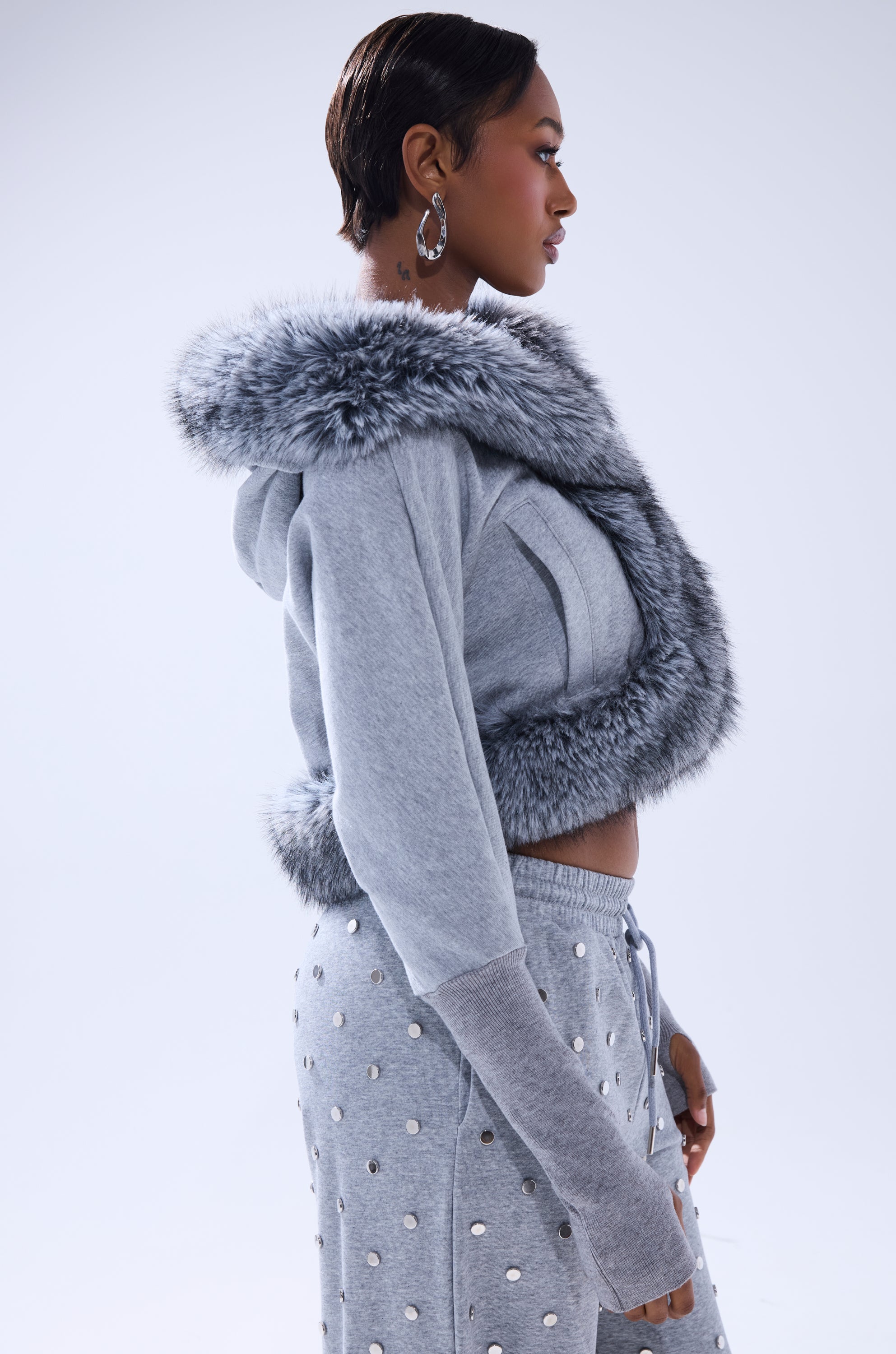 A person in profile models the MOVIN DIFFERENT SWEATSHIRT WITH FUR TRIM in light gray, featuring faux fur on the hood and hem, paired with matching metallic silver polka dot sweatpants against a plain light background. 5