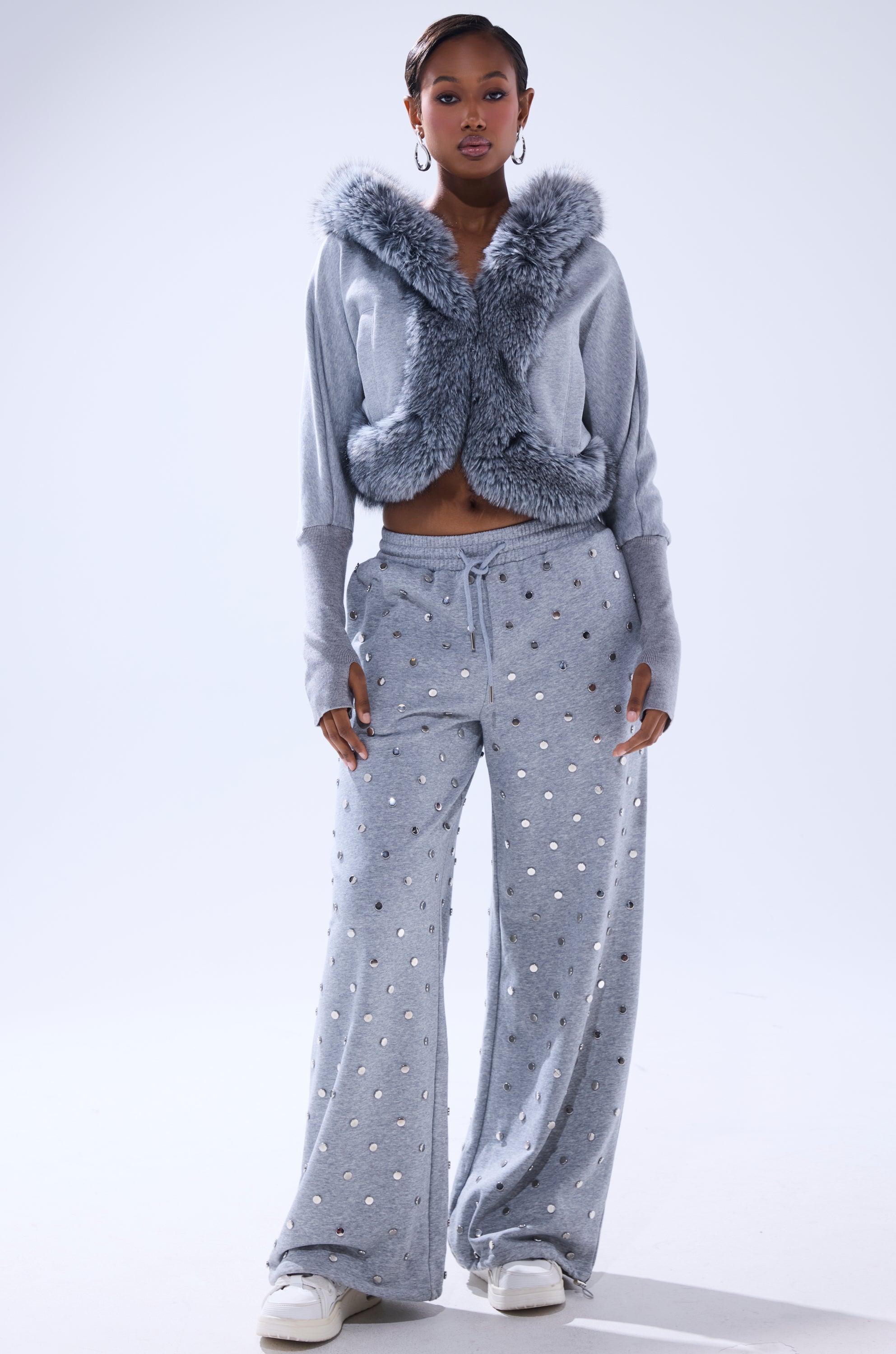 A woman faces forward in the MOVIN DIFFERENT SWEATSHIRT WITH FUR TRIM, a light gray cropped faux fur top, paired with wide-leg silver polka dot pants, white sneakers, and hoop earrings for casual luxury. 7