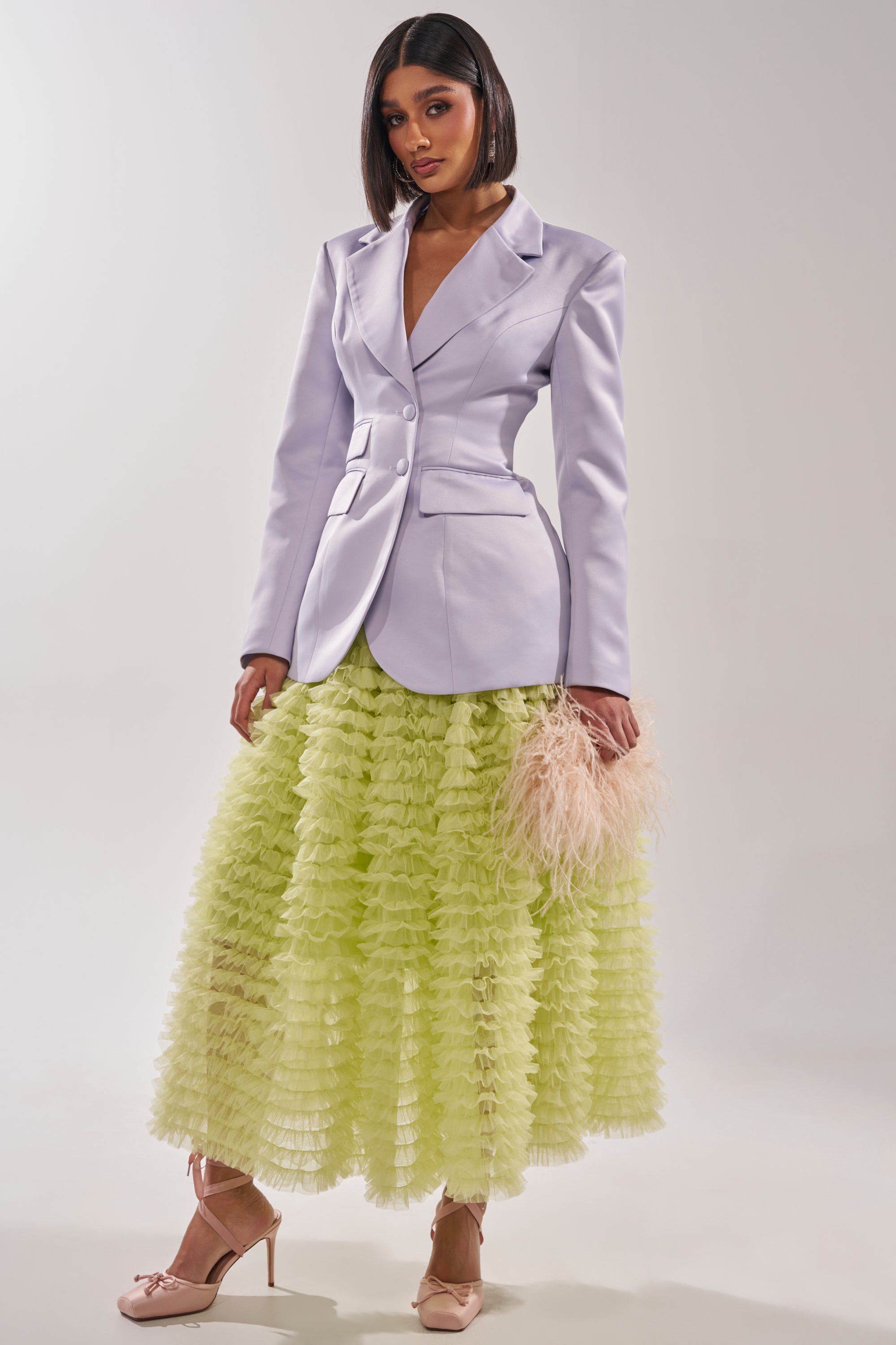 A woman models the PERFECT FIT BLAZER IN LILAC styled with a long, ruffled pastel yellow skirt, a pale pink feathered clutch, and nude heels against a simple light background. 10