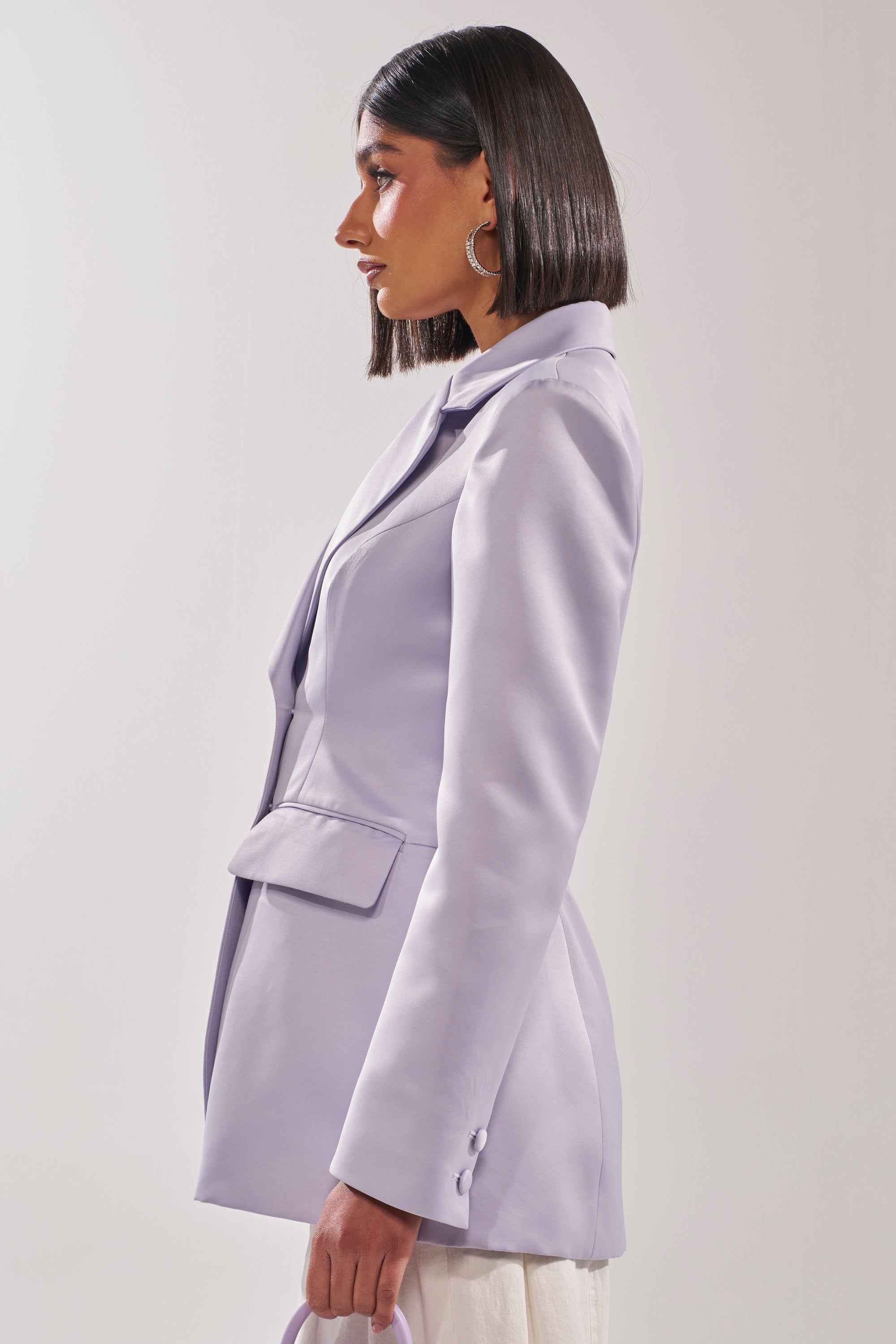 A woman with straight, shoulder-length hair is seen in profile against a plain light background, wearing the PERFECT FIT BLAZER IN LILAC and holding a matching bag. 2