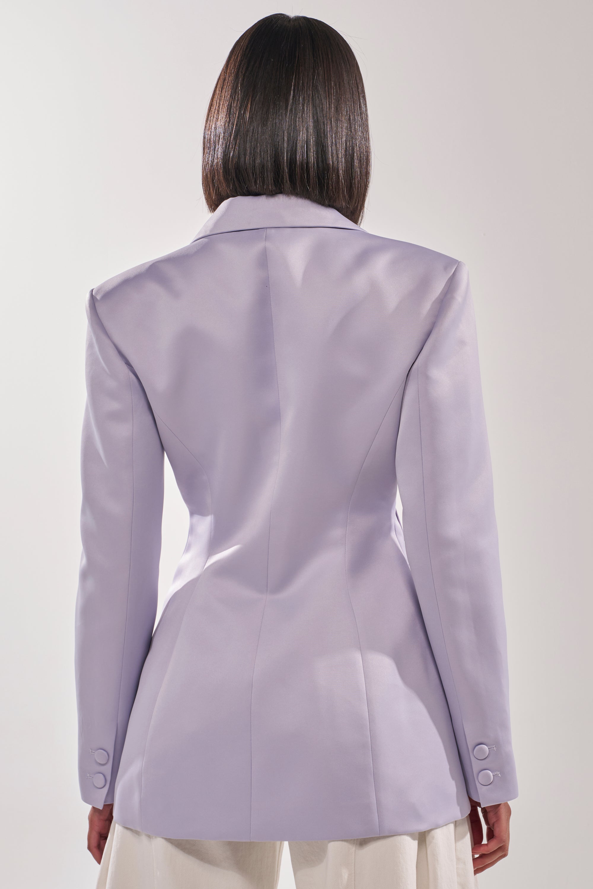 A person with straight, shoulder-length dark hair is seen from the back wearing the PERFECT FIT BLAZER IN LILAC, a tailored longline women's office blazer, paired with white pants against a plain light background. 3