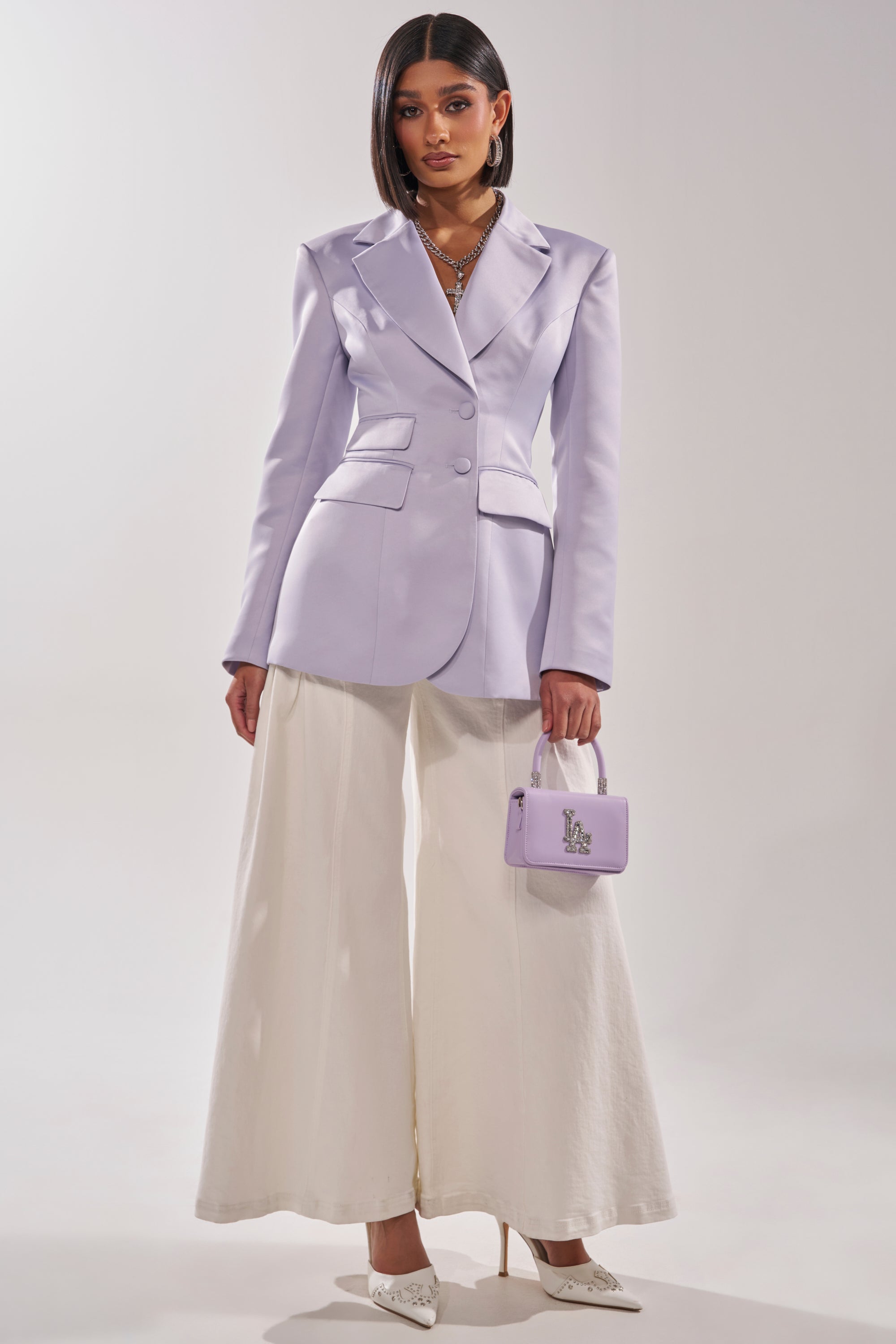 A woman wears the PERFECT FIT BLAZER IN LILAC, styled with wide-leg white trousers, pointed white heels, a matching lilac handbag, layered necklaces, and her straight shoulder-length hair completes the look. 4