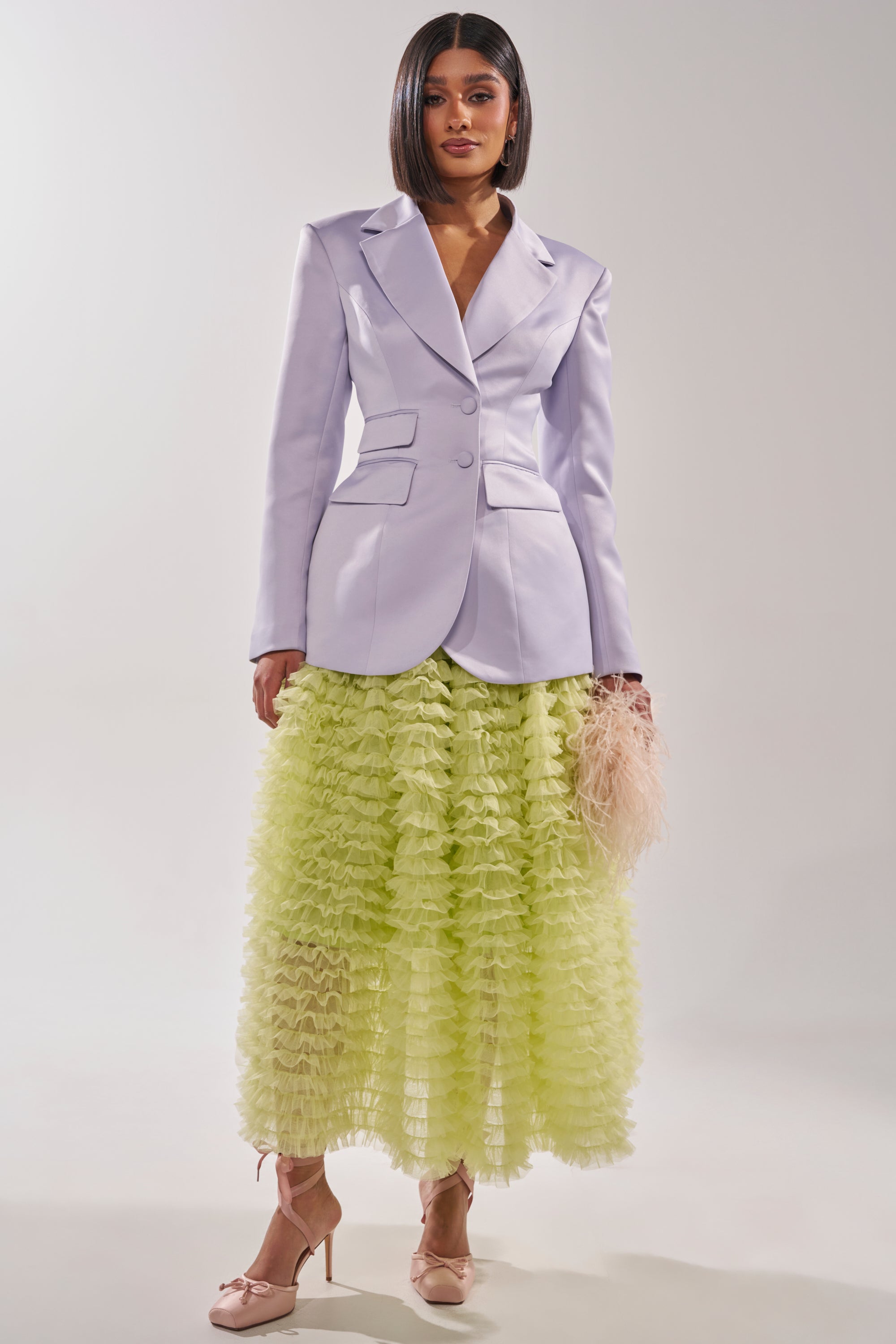 A woman stands against a plain light background wearing the PERFECT FIT BLAZER IN LILAC, a yellow ruffled skirt, nude heels, and holding a feathery pale pink clutch. 6