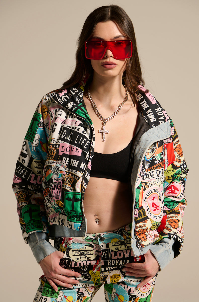 A woman stands confidently in the GREEN LIGHT LAYERED GRAPHIC BOMBER with matching pants, a black crop top, red oversized sunglasses, and a silver cross necklace against a neutral background.