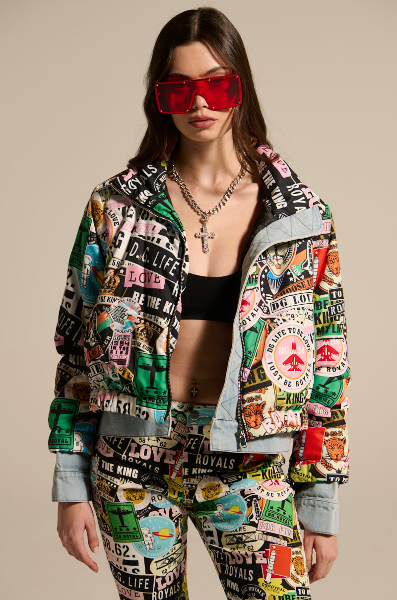 A woman wearing the GREEN LIGHT LAYERED GRAPHIC BOMBER over a black crop top, vibrant graphic patchwork street-style pants, oversized red sunglasses, and a chunky chain necklace with a cross pendant.