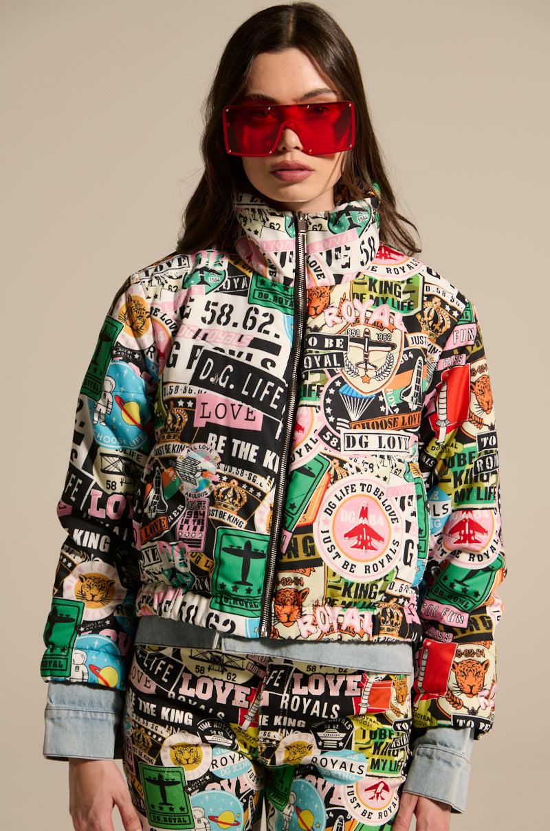 A woman wearing the GREEN LIGHT LAYERED GRAPHIC BOMBER—a colorful, street-style patchwork jacket with bold graphic text and images—stands against a neutral background.