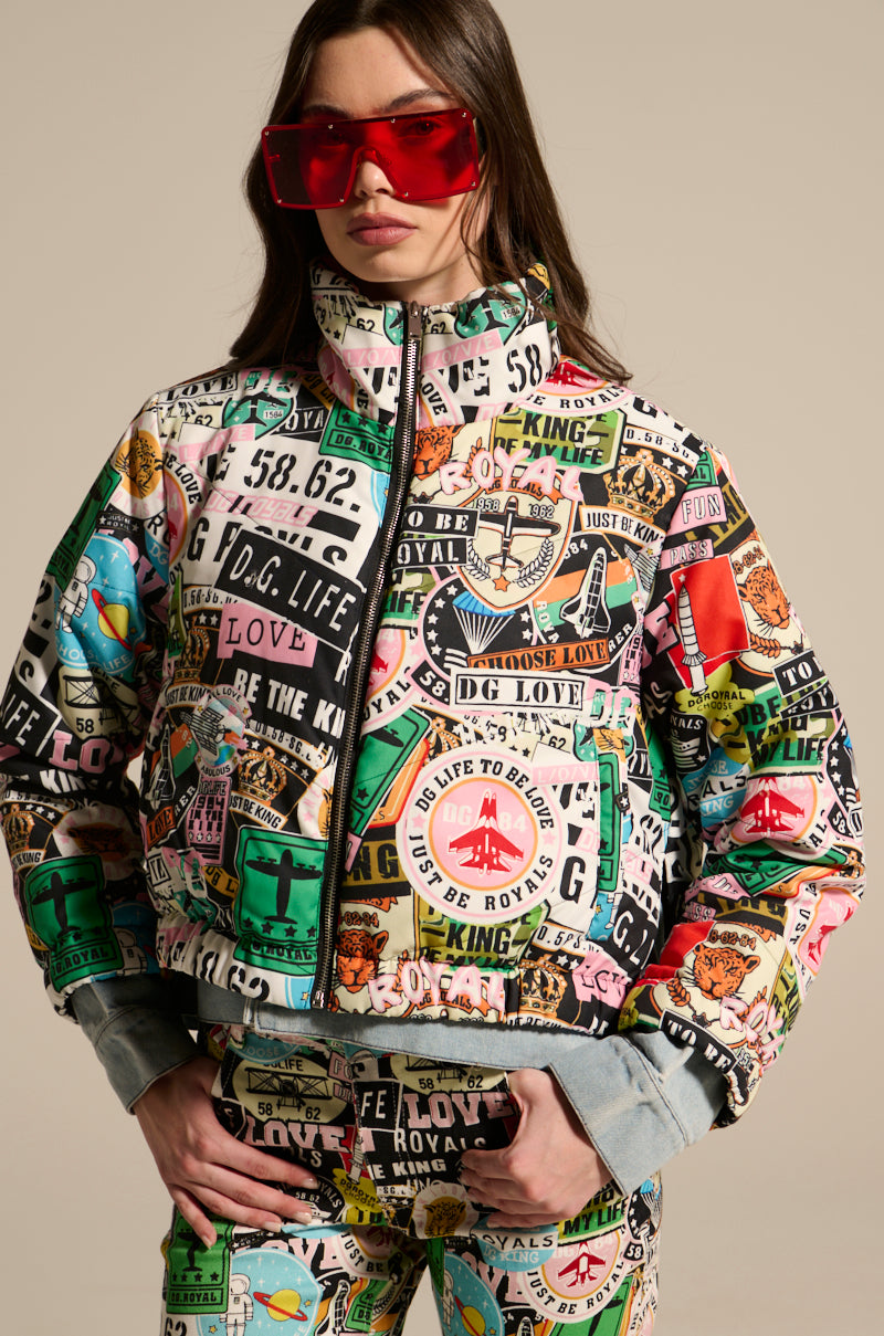 A woman wearing the GREEN LIGHT LAYERED GRAPHIC BOMBER, styled with oversized red sunglasses and graphic patch pants, stands confidently against a neutral background.