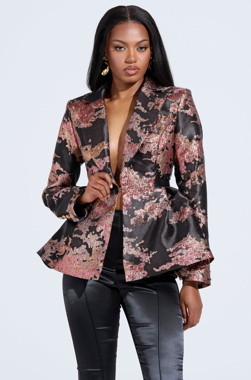 A person wearing the CANARY PEPLUM BLAZER IN BLACK MULTI with a deep neckline and black satin pants poses confidently. They have long, dark hair and gold hoop earrings, standing against a plain light gray background. 0