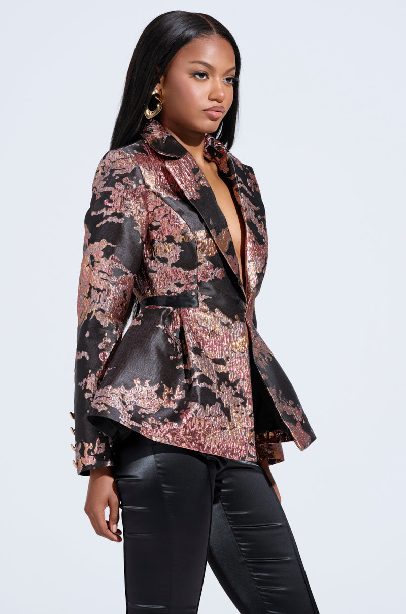 A woman in a chic floral brocade CANARY PEPLUM BLAZER IN BLACK MULTI with gold buttons and black leather pants stands against a neutral backdrop, gazing sideways. She sports long black hair and gold hoop earrings. 1