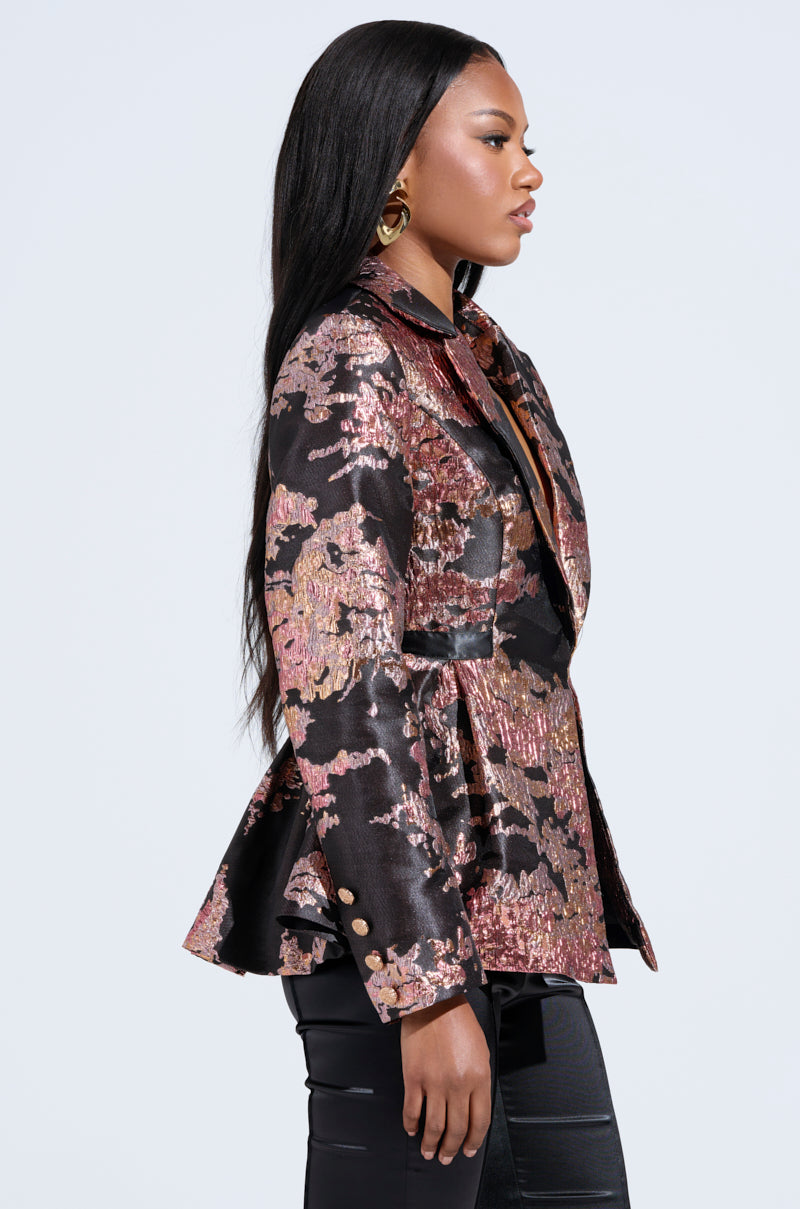 A woman with long dark hair stands in profile, wearing the CANARY PEPLUM BLAZER IN BLACK MULTI with metallic threads and gold buttons, paired with black pants. She features gold earrings and a confident expression against a plain white background. 2