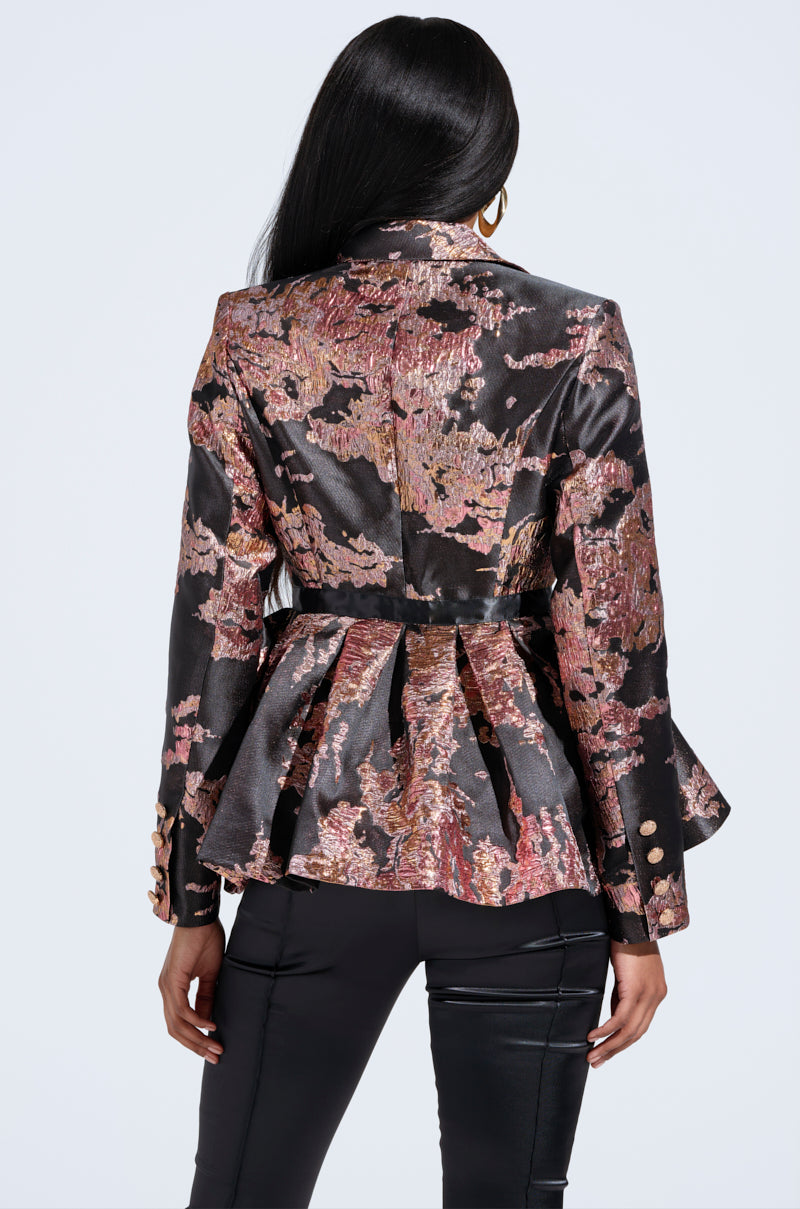 Rear view of a person with long dark hair wearing the CANARY PEPLUM BLAZER IN BLACK MULTI, featuring a metallic texture with rich pink and gold hues, gold buttons, intricate floral brocade on the sleeves, flared waist styling, and paired with dark pants. 3