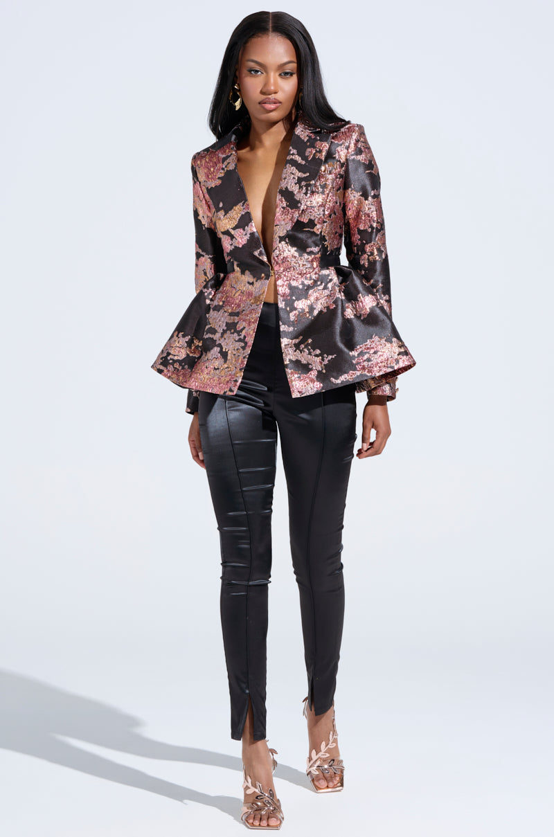 Against a plain white background, a woman poses in the CANARY PEPLUM BLAZER IN BLACK MULTI, featuring a floral brocade pattern with a deep neckline and flared hem. She pairs it with black leather pants with front slits and elegant heels, her long hair straight. 5