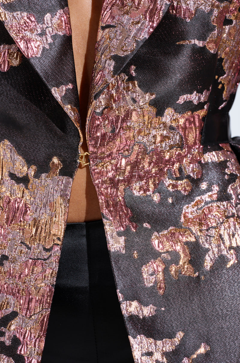 A close-up features a person in the CANARY PEPLUM BLAZER IN BLACK MULTI, showcasing its metallic gold, pink, and black abstract patterns. The textured satin blazer is partly fastened with shiny gold buttons, revealing a hint of the chest. 6