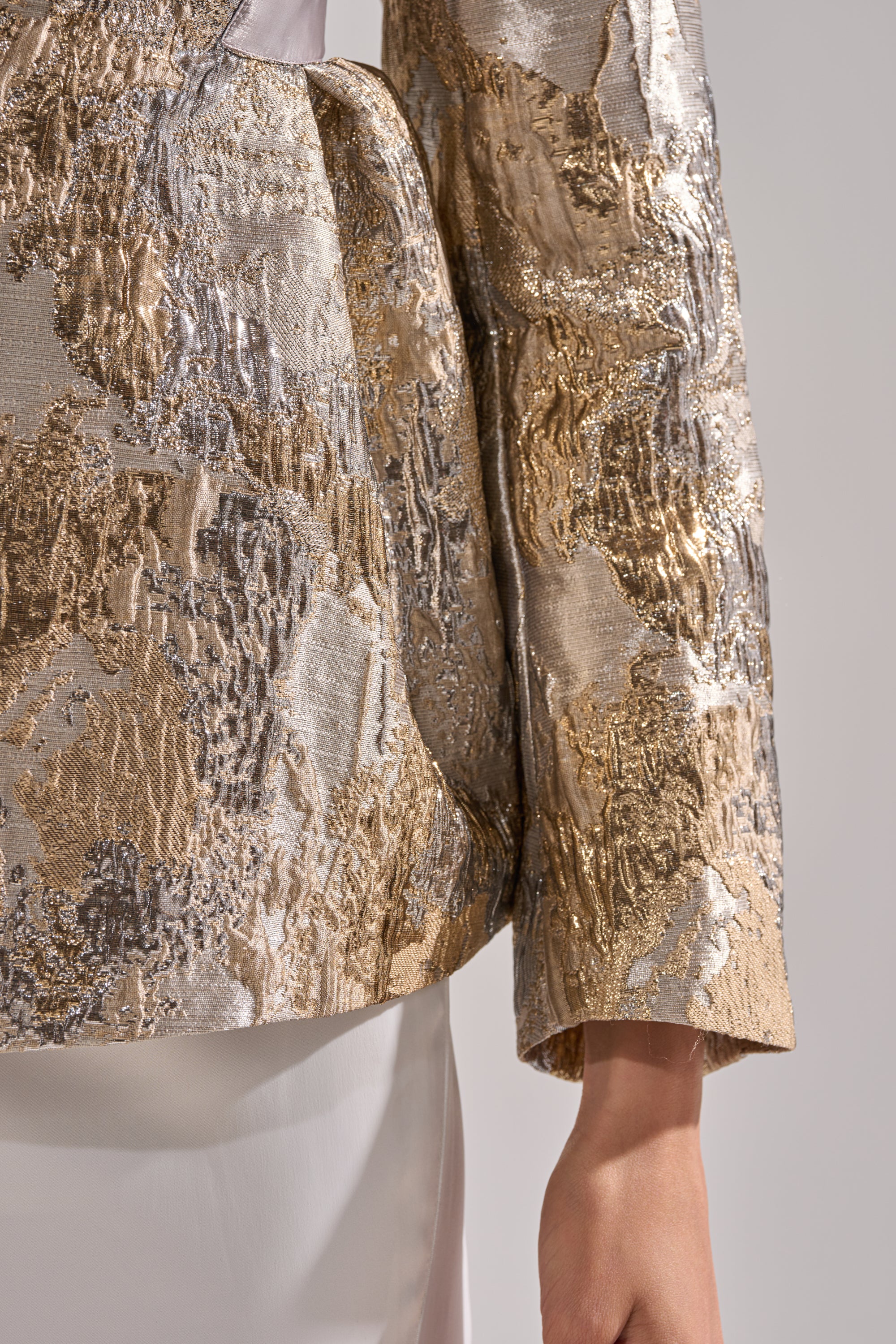 A close-up shows the lower sleeve, waist, and hand of a person wearing the CANARY PEPLUM BLAZER IN CHAMPAGNE over a white outfit, set against a neutral background.
