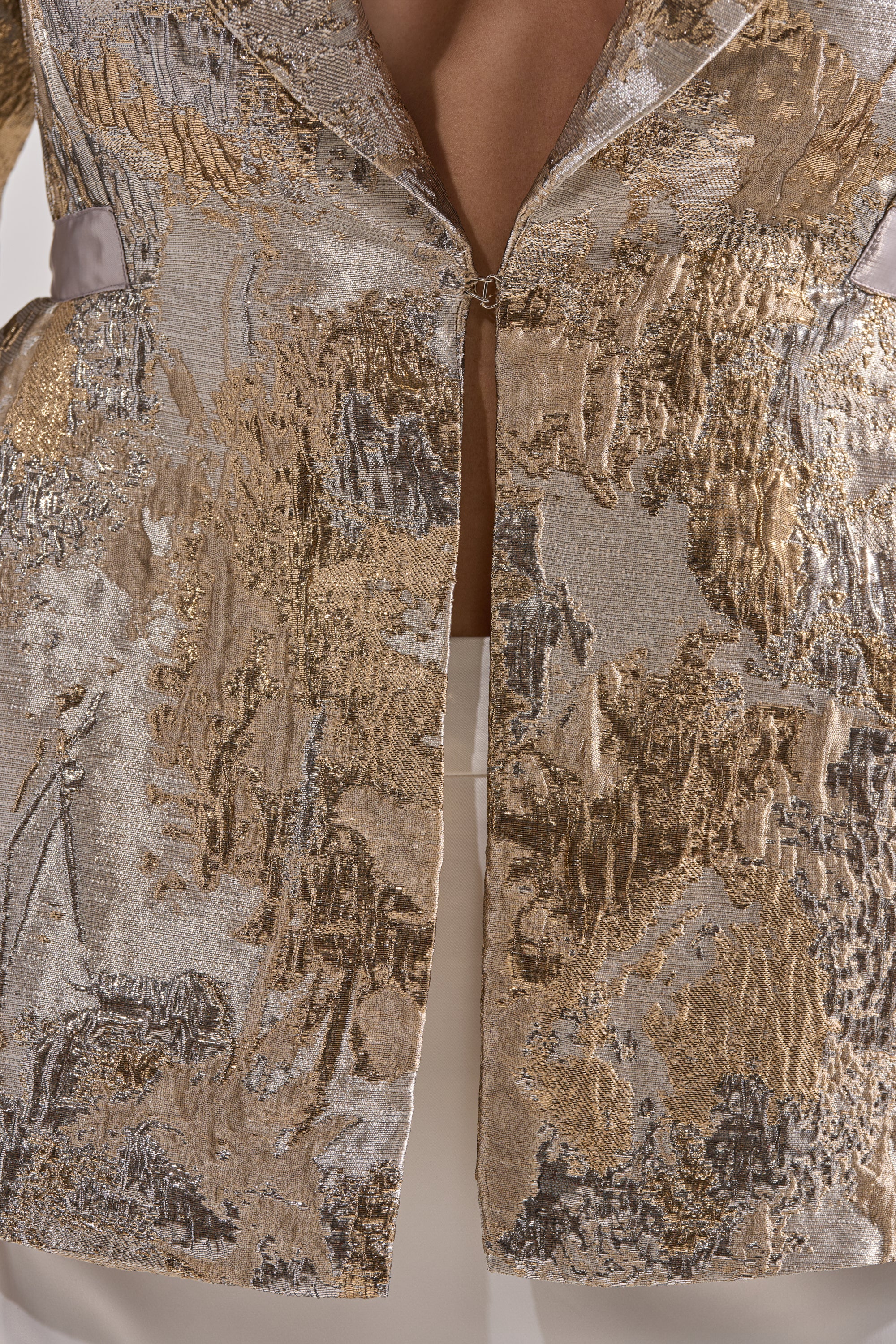 A close-up of the CANARY PEPLUM BLAZER IN CHAMPAGNE, featuring metallic brocade with gold and silver patterns, worn over white pants. The partially open blazer reveals a hint of skin underneath.