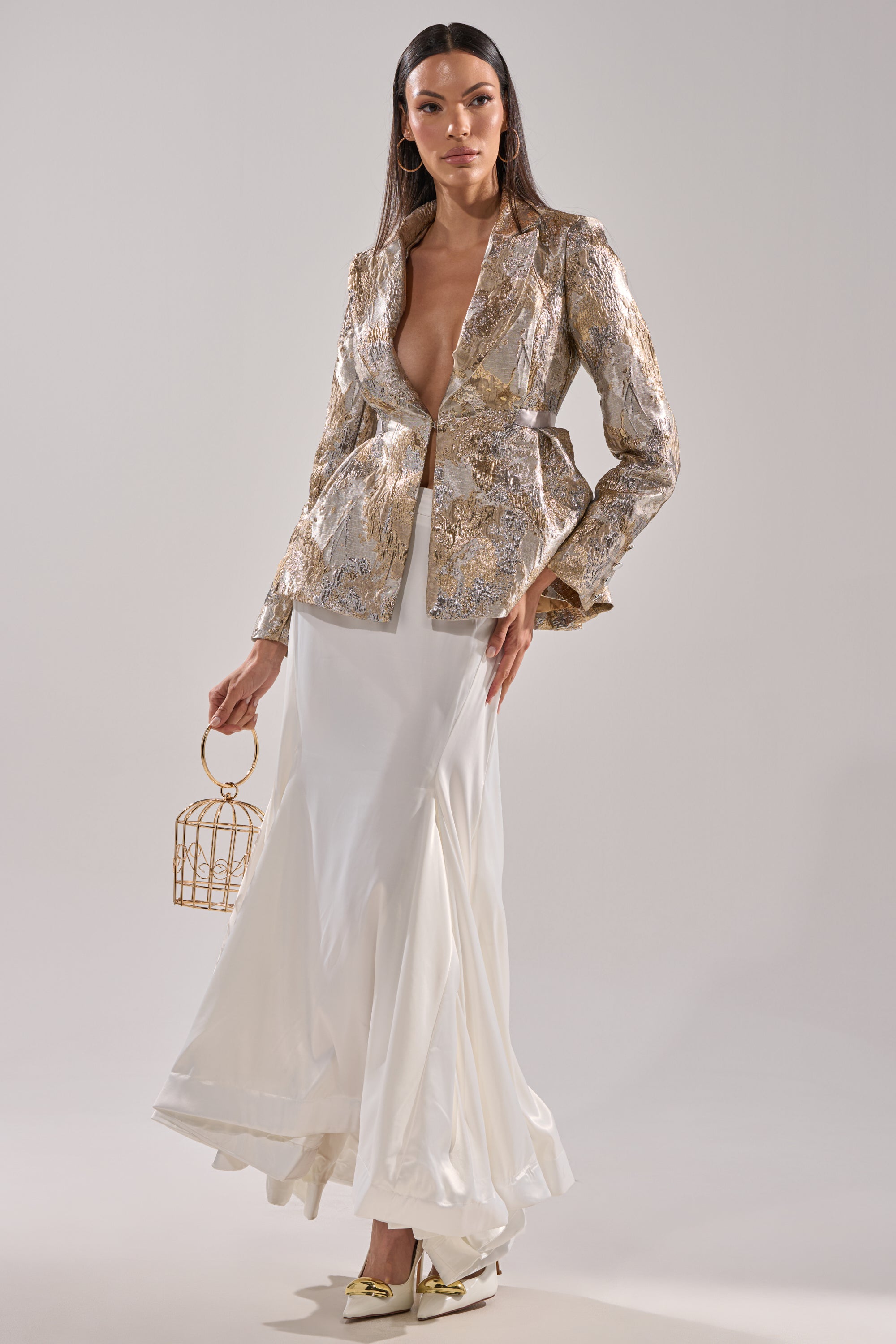 A woman in the CANARY PEPLUM BLAZER IN CHAMPAGNE over a white flowing skirt holds a gold birdcage handbag. She has straight dark hair, gold heels, and stands against a plain light background.