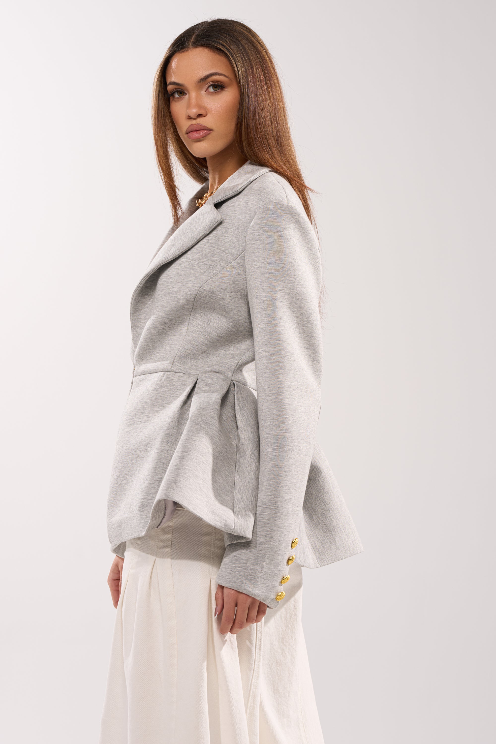 A woman with long, straight brown hair wears the CANARY PEPLUM BLAZER IN SCUBA in light gray with gold buttons and a flared hem over a flowing white skirt, creating an elegant hourglass silhouette against a plain light background. 2