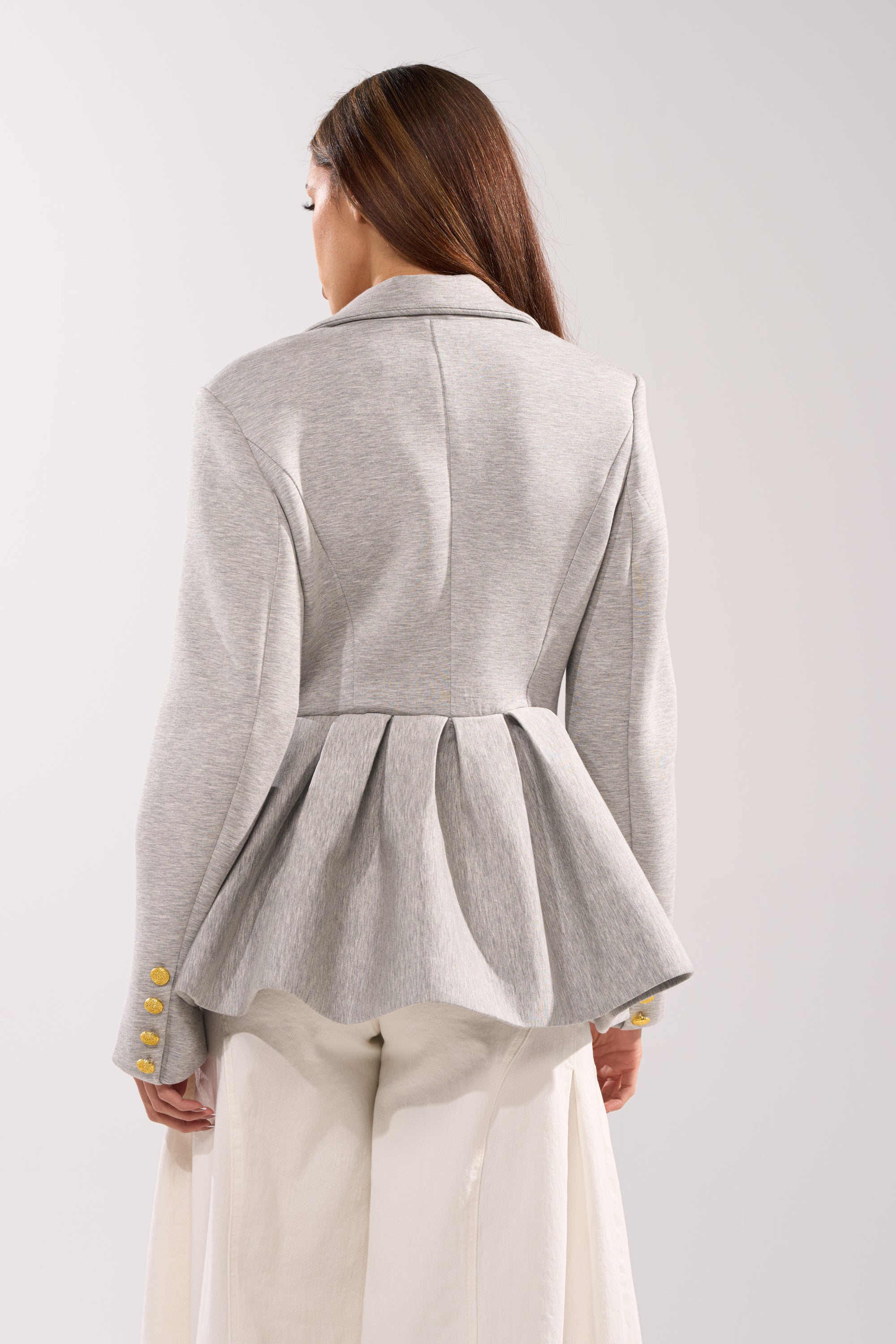 A woman with long brown hair wears the CANARY PEPLUM BLAZER IN SCUBA in light gray, tailored for an hourglass silhouette with gold buttons, paired with wide-leg white trousers, against a plain light background. 3