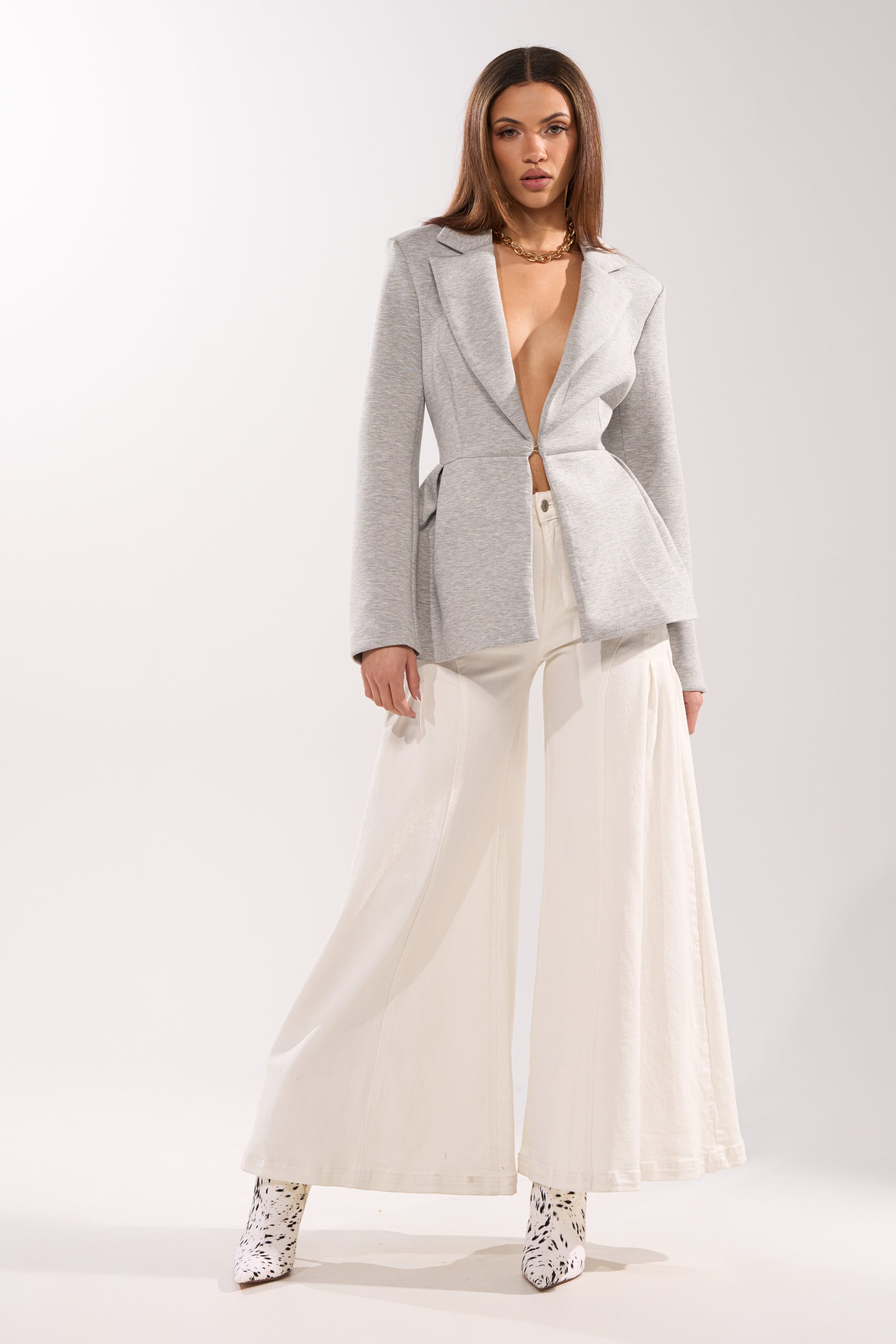A woman poses against a plain background in the CANARY PEPLUM BLAZER IN SCUBA, paired with wide-legged white pants, patterned white ankle boots, and a chunky gold necklace. 4