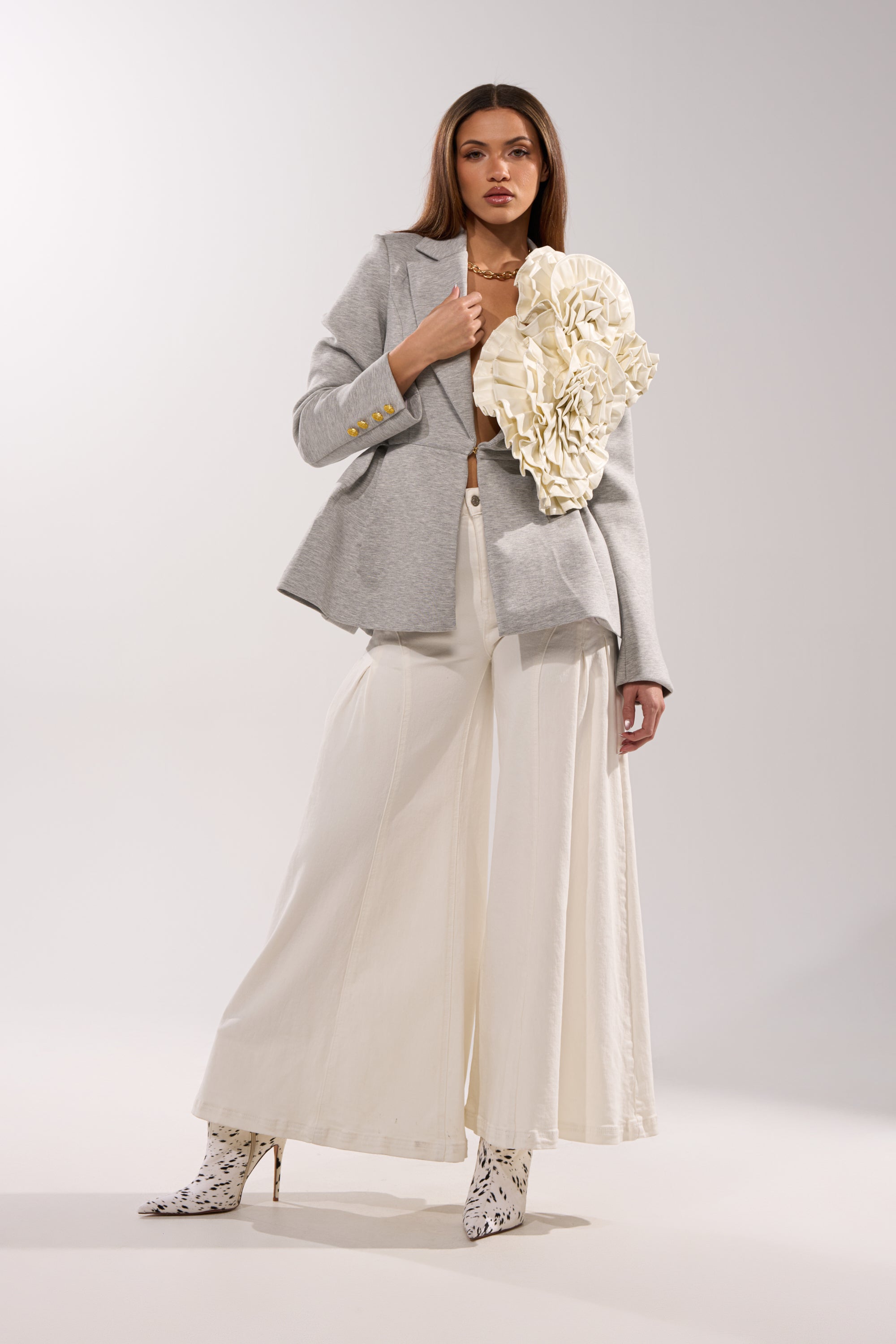 A woman stands confidently in the CANARY PEPLUM BLAZER IN SCUBA, paired with wide-leg white pants and white heeled boots with black patterns, set against a plain background. 6