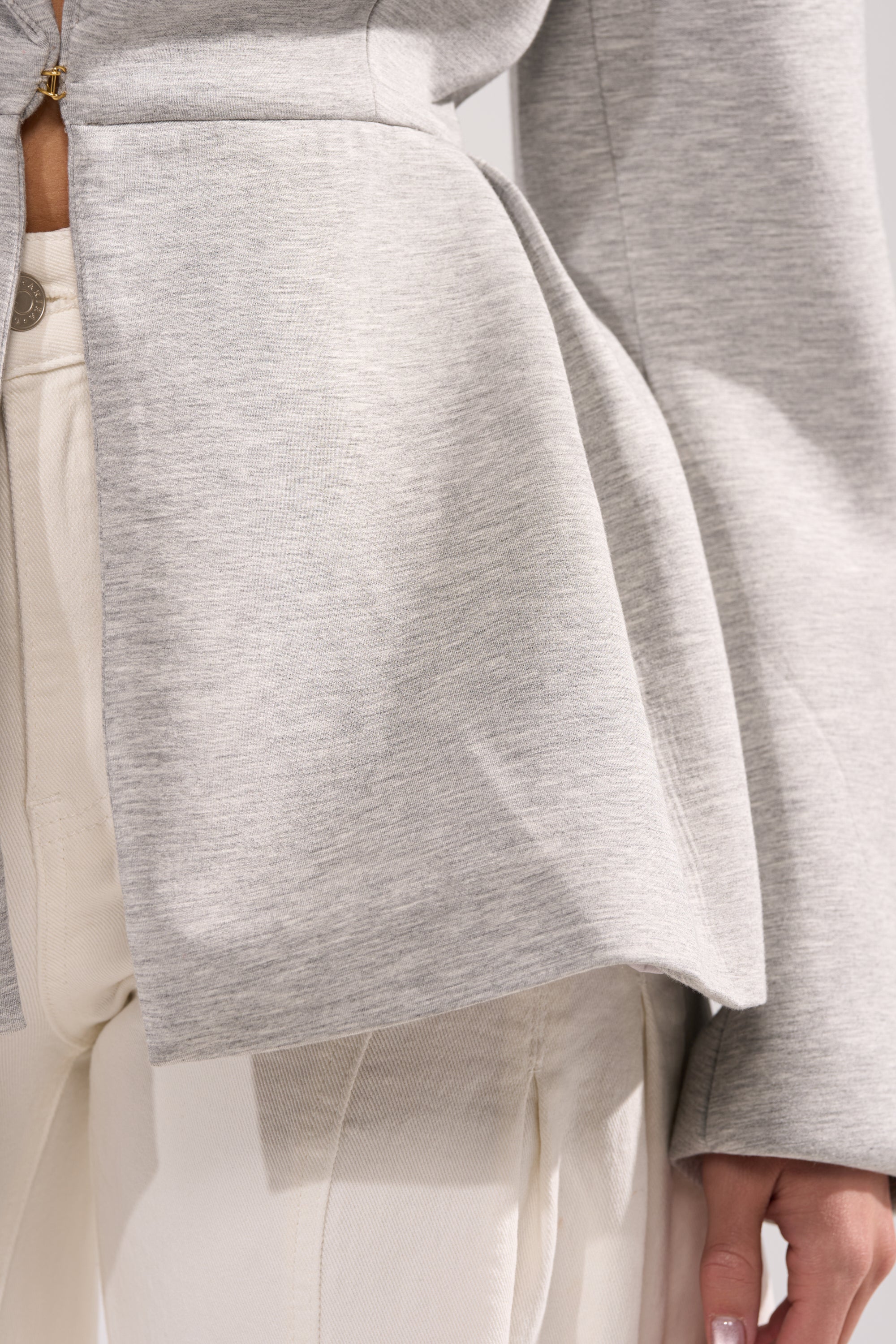 A close-up shows a person wearing the CANARY PEPLUM BLAZER IN SCUBA in light gray over cream pants, highlighting its textured scuba stretch fabric and showing part of the person's hand. 7