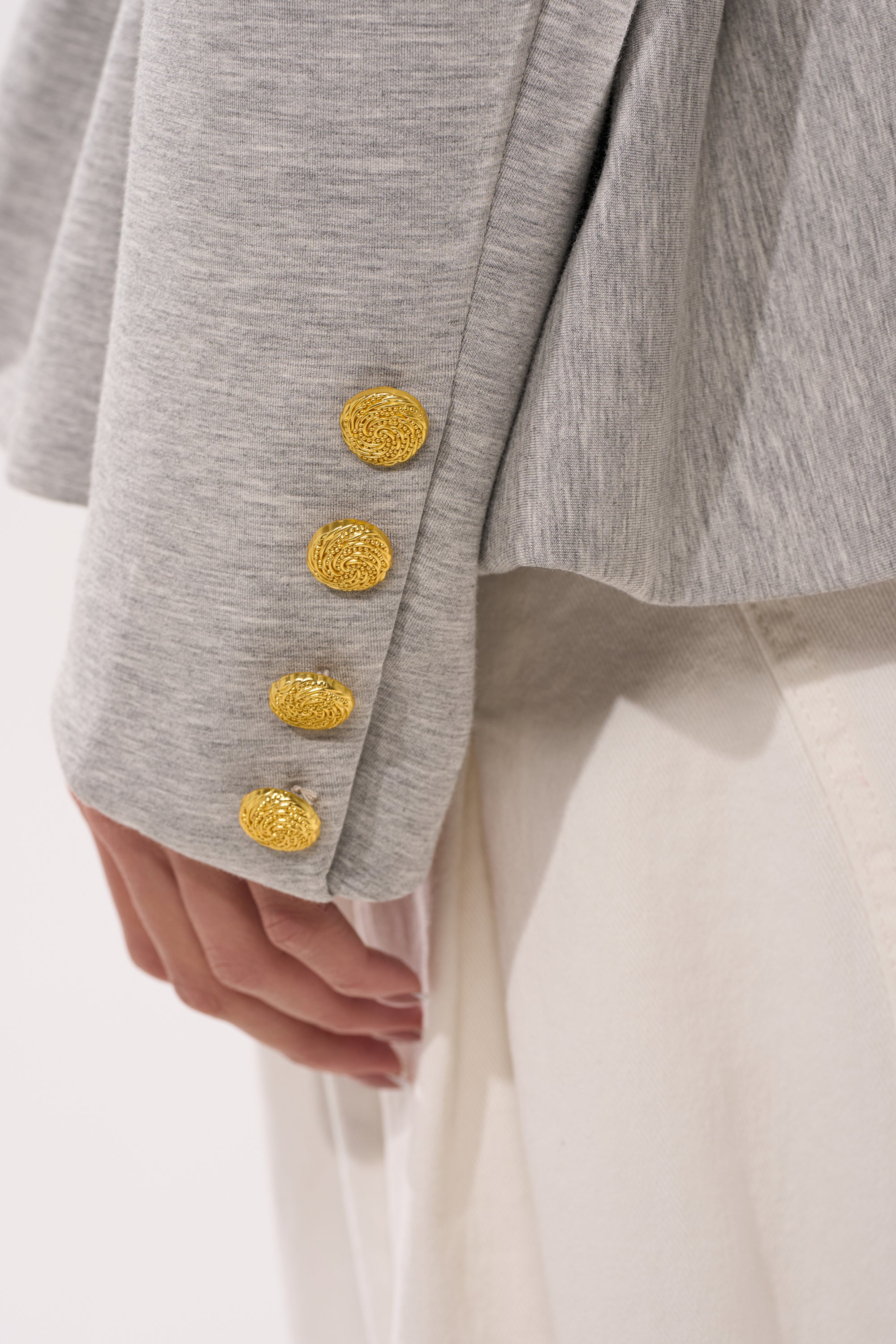 A close-up of a person’s arm in the CANARY PEPLUM BLAZER IN SCUBA, featuring light gray fabric and four textured gold cuff buttons, paired with white pants. The hourglass silhouette stands out as their hand rests at their side. 8
